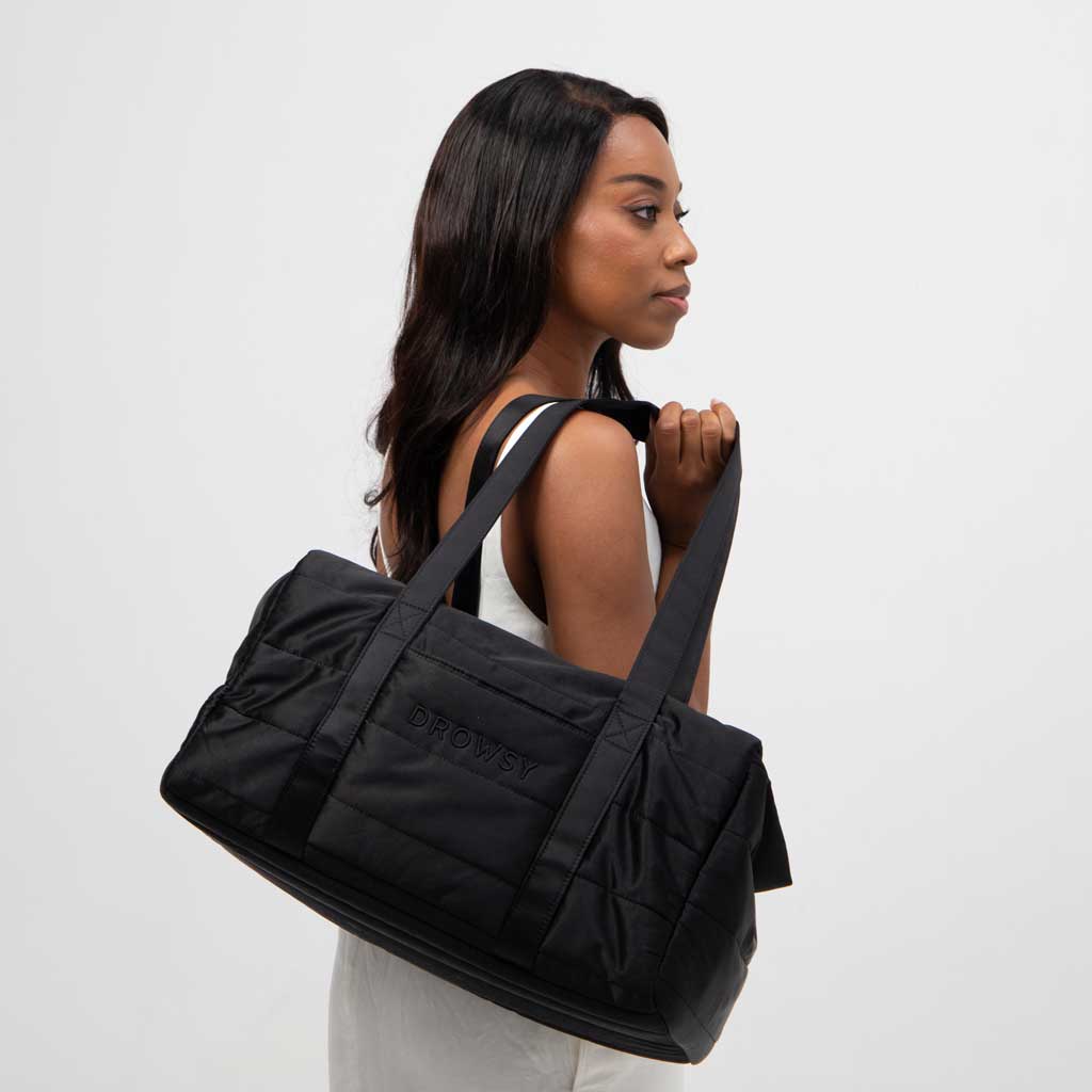 A woman with long dark hair in a white sleeveless top holds the Drowsy Silk Weekender - Black Jade duffel bag by Drowsy, featuring the brand name on the side, standing against a plain white background.