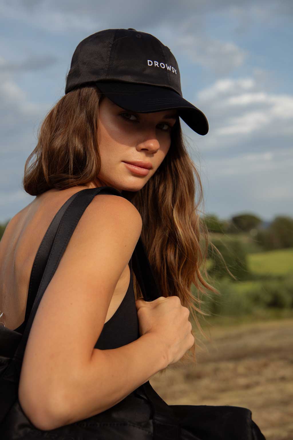 A woman with long brown hair wearing a black cap and sleeveless top looks over her shoulder outdoors, holding a backpack strap, with green fields and a cloudy sky in the background.