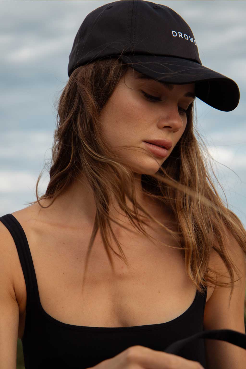 A woman with long brown hair wearing a black cap and black tank top looks down with a calm expression. The sky is cloudy in the background.
