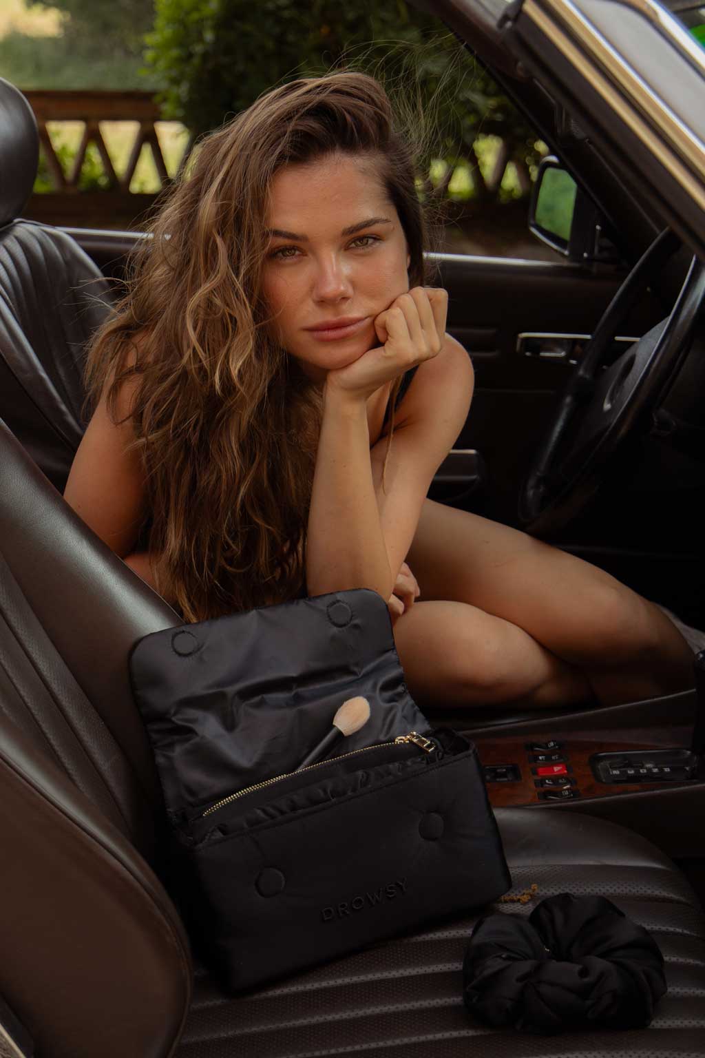 A woman with long wavy hair sits in a car, resting her chin on her hand. An open black makeup bag with a brush inside and a scrunchie are on the car seat next to her.