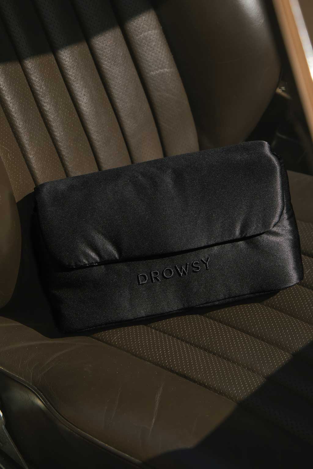 A black pouch with the word DROWSY embroidered on it sits on a brown, leather car seat in sunlight.