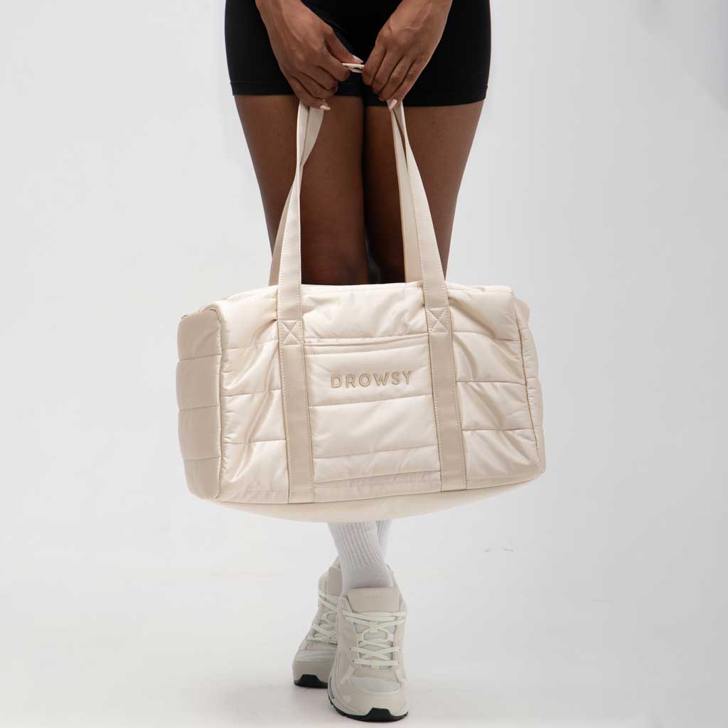 A person holds the Drowsy Silk Weekender in Dusty Gold—a chic, quilted duffle bag by Drowsy—against a plain, light background while wearing black shorts, white socks, and white sneakers.
