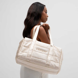 A woman with long dark hair carries the Drowsy Silk Weekender in Dusty Gold, featuring quilted cream-colored silk and the DROWSY logo embroidered on the side, set against a plain light background.