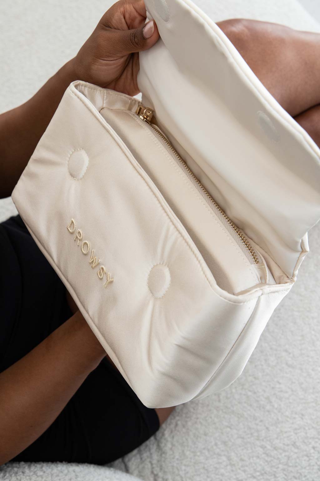 A person holds an open, cream-colored, padded pouch with the word DROWSY embroidered on it in gold letters. The pouch has a zippered compartment and is held on the persons lap.