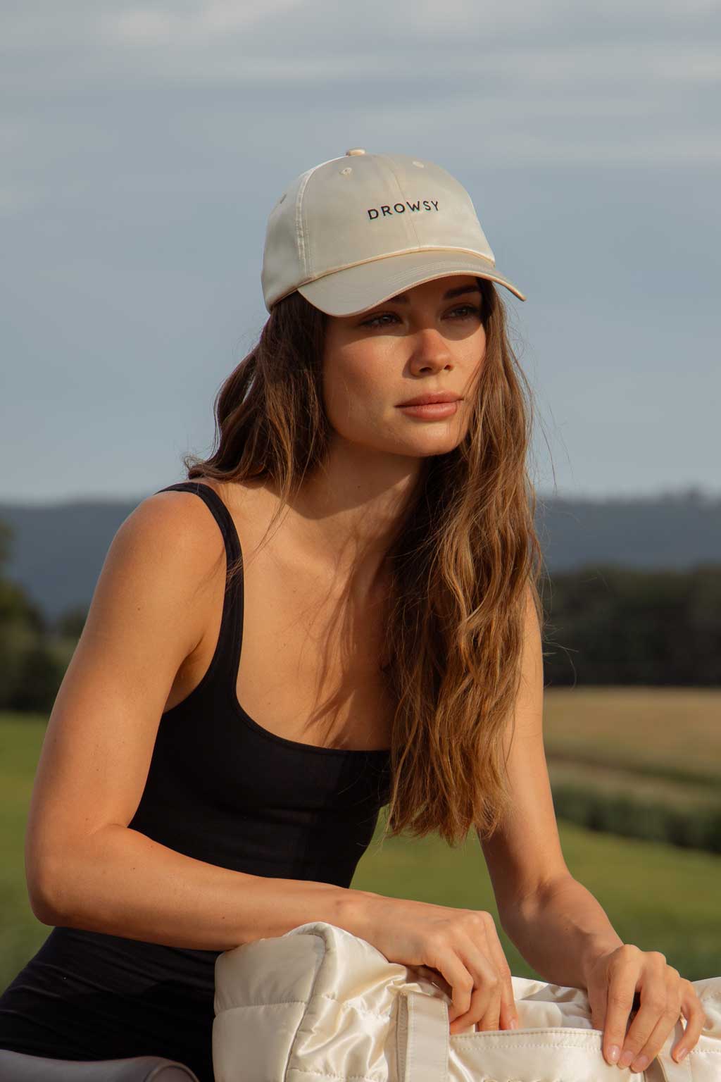 A woman with long brown hair wearing a light-colored Drowsy cap and a black tank top sits outdoors in a grassy field, holding a light-colored bag. Trees and hills are visible in the background.