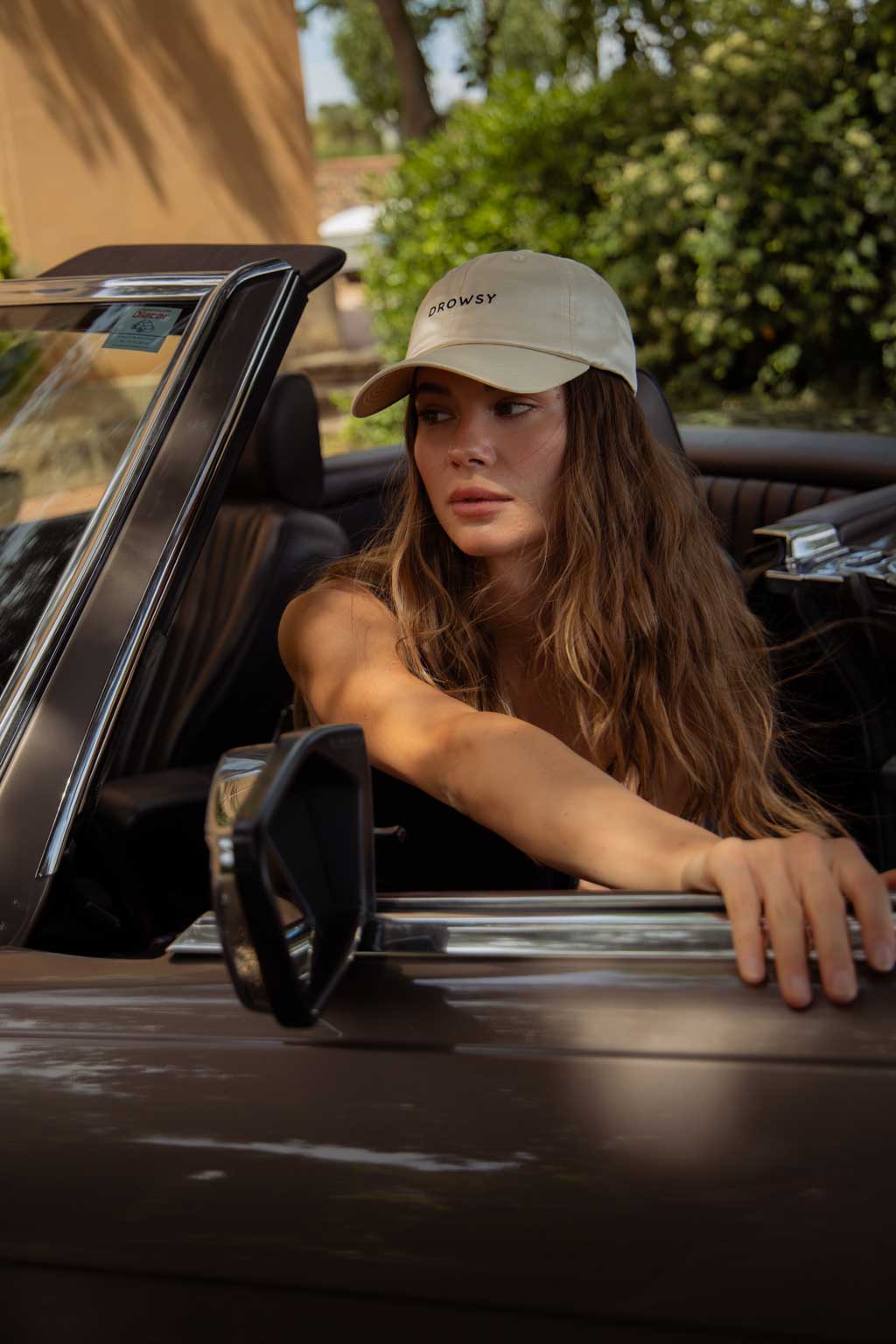 A woman with long wavy hair, wearing a beige cap and black top, sits in the driver’s seat of a convertible car with her left hand on the steering wheel and right arm resting on the door, looking thoughtfully to her right.
