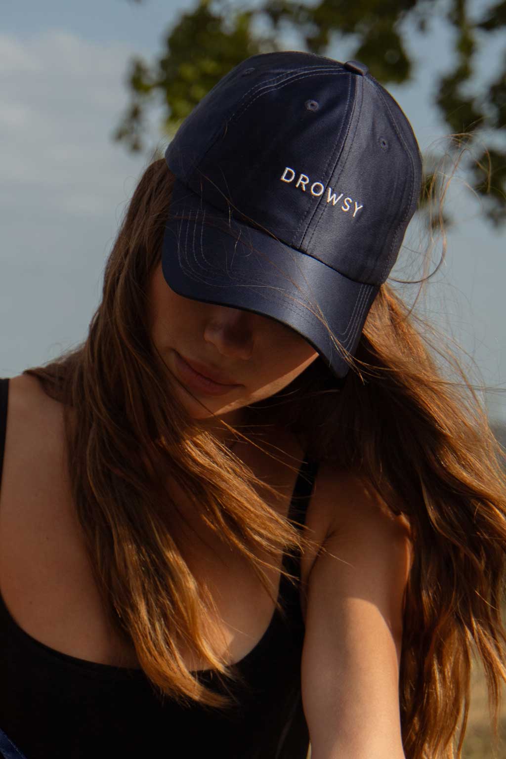 A person with long brown hair wearing a navy blue cap with the word “DROWSY” embroidered on the front, looking downward, outdoors with blurred trees and sky in the background.