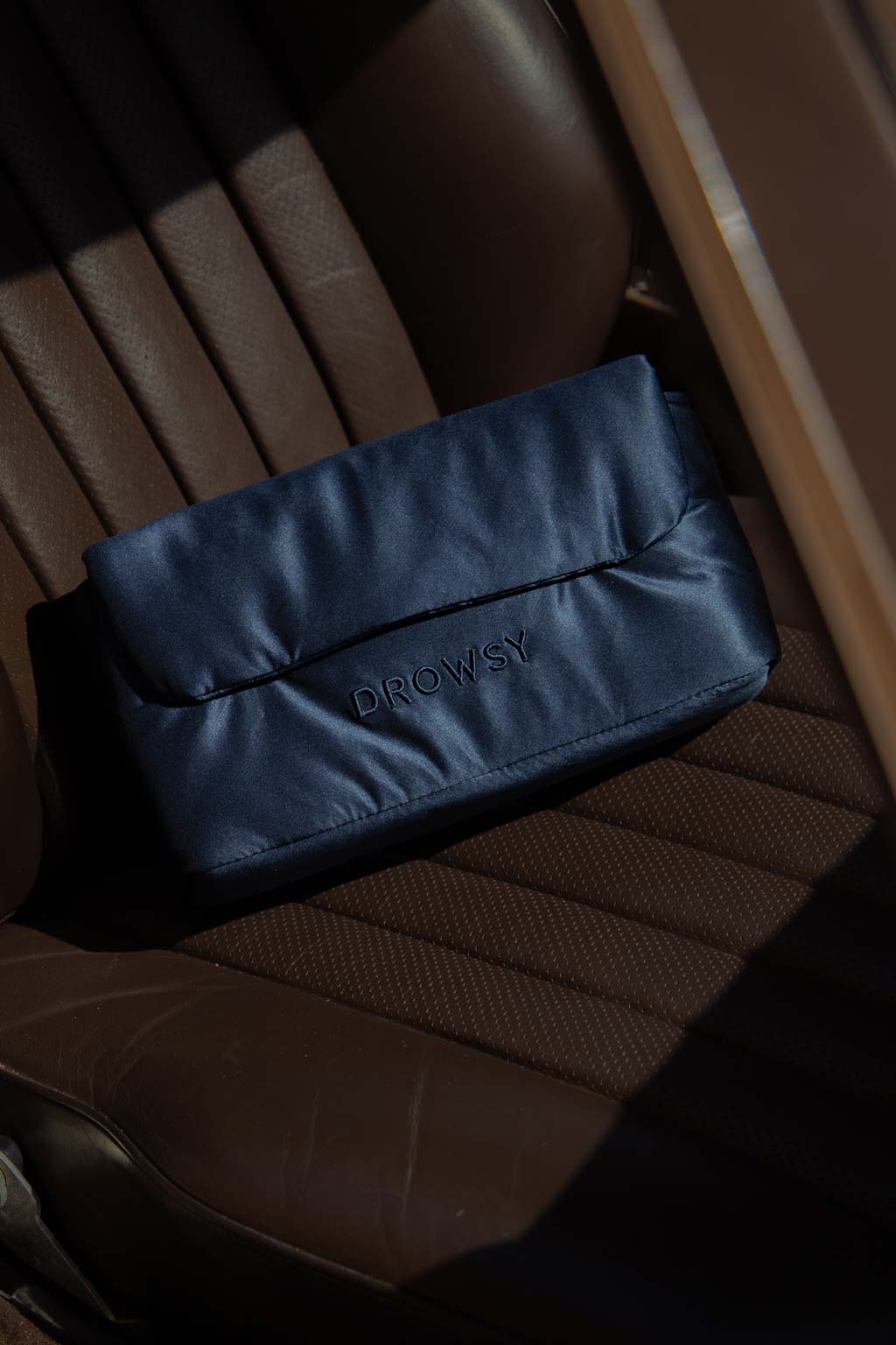 A navy blue pouch with the word DROWSY embroidered on it sits on a brown leather car seat, partially in shadow.