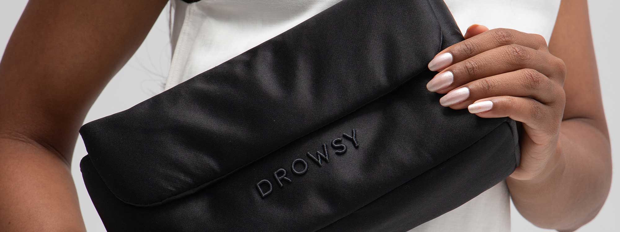 A person with neatly manicured nails holds a black pouch with DROWSY embroidered on it, against a white top.