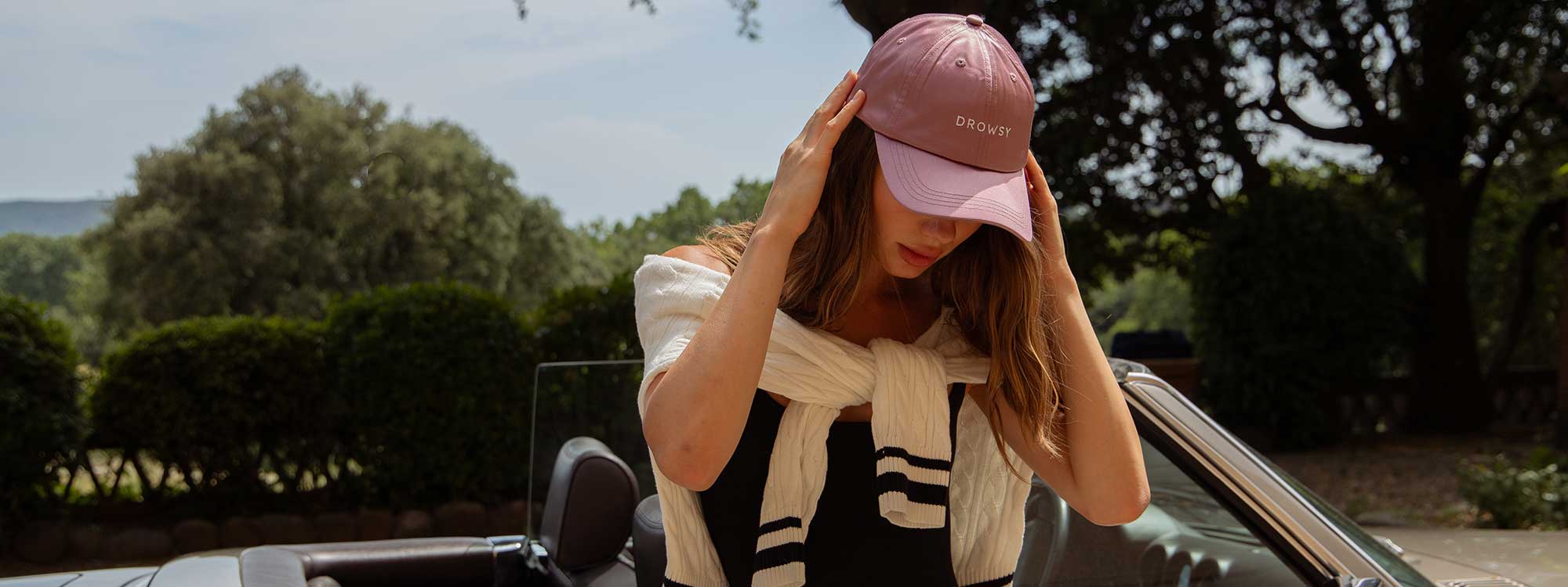 A woman wearing a pink cap and black outfit stands next to a convertible car, adjusting her hat. She has a white sweater draped over her shoulders. Lush green trees and a sunny sky are in the background.