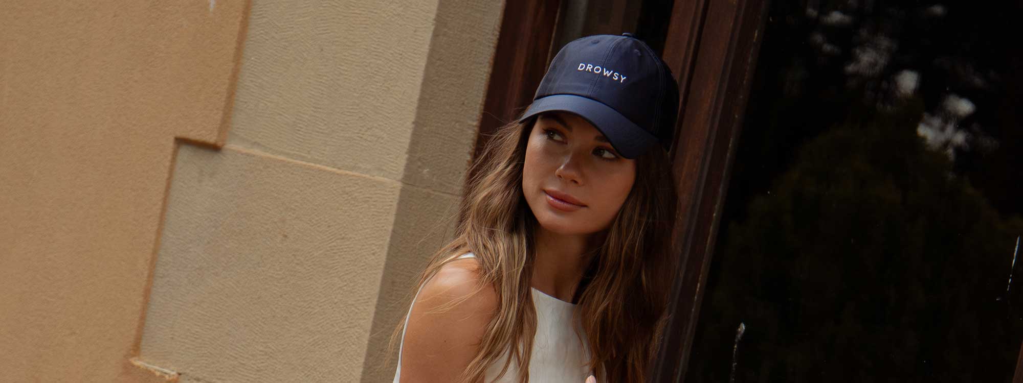 A woman with long brown hair wearing a navy blue cap and a white sleeveless top stands outdoors near a stone wall, looking to the side.
