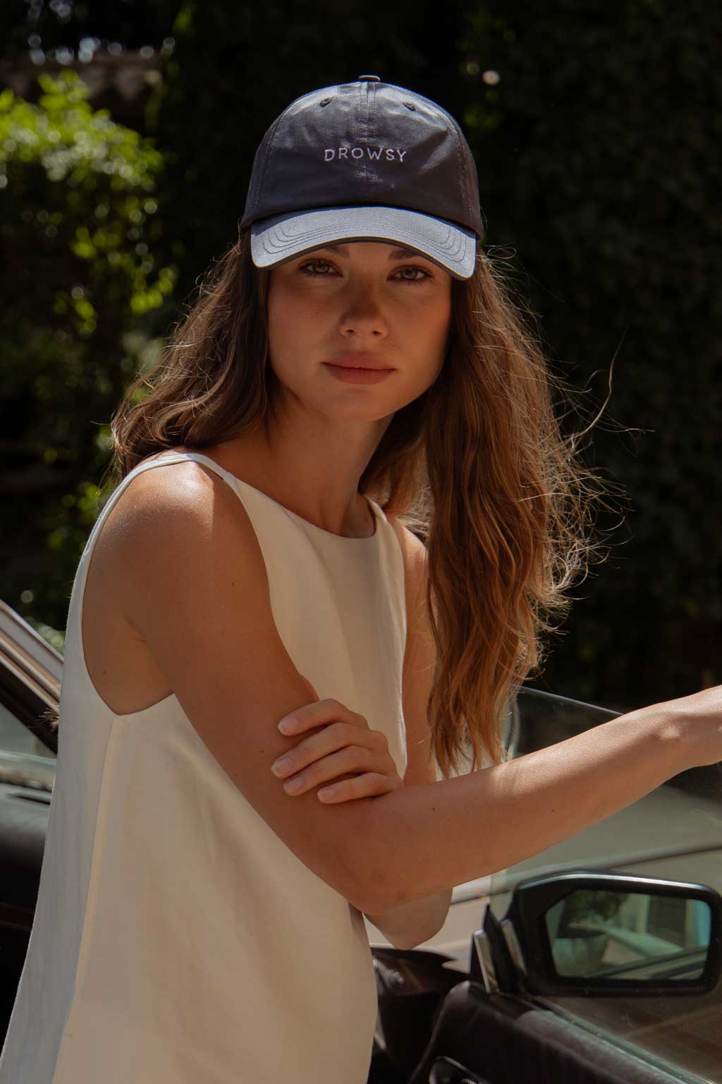 A woman with long wavy hair wearing a sleeveless white top and a navy blue cap that says DROWSY stands next to a car door, looking at the camera, with greenery in the background.