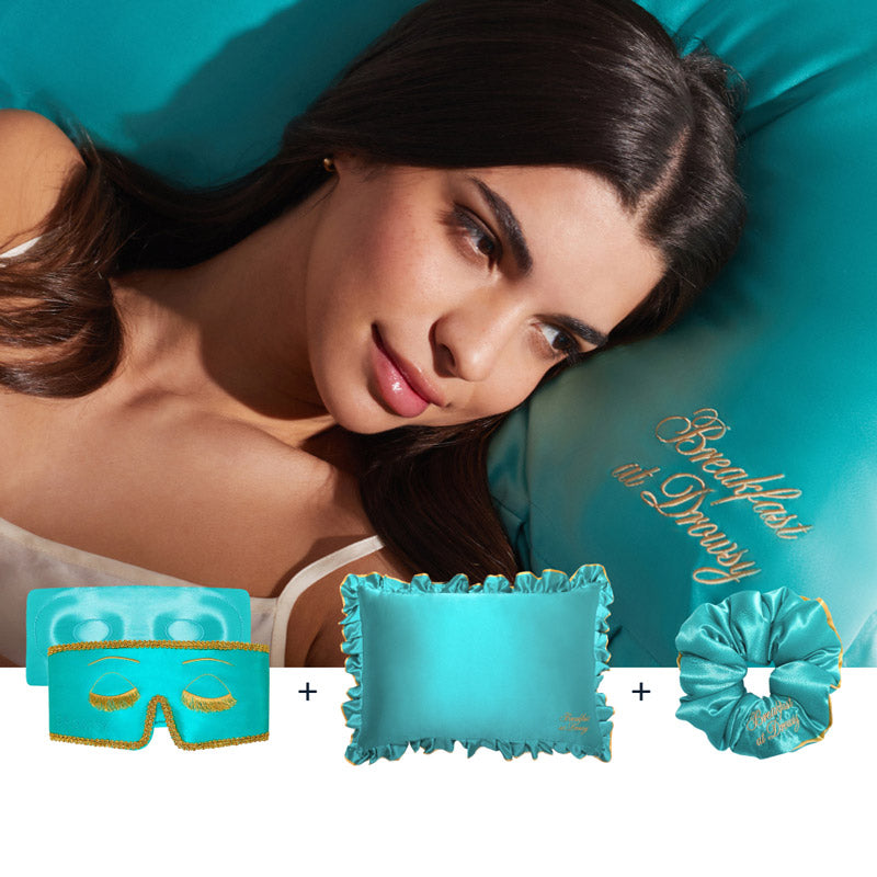 A woman with long dark hair relaxes on a teal satin pillow from the Hollywood Experience Bundle, featuring a silk sleep mask, matching pillow, and scrunchie—all with gold trim for the perfect beauty sleep set.