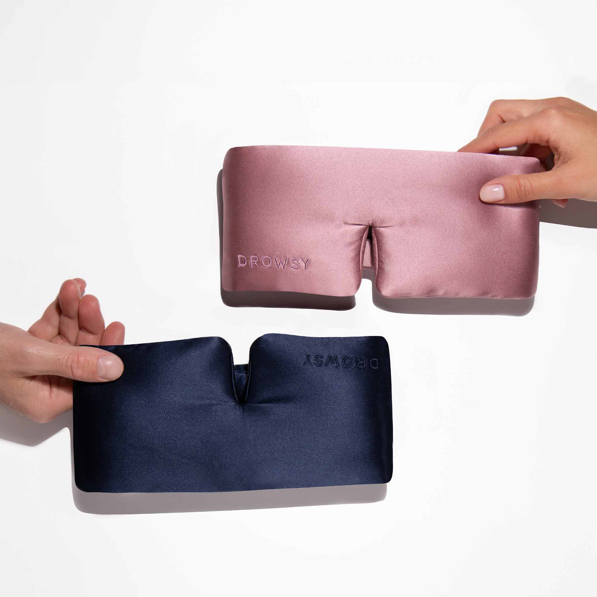 Two hands each hold a silky sleep mask labeled Drowsy; one mask is pink, the other is dark blue, both displayed against a white background.
