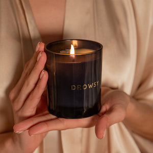 A person in a light robe gently holds a lit Sleep Ritual Candle 220g in a dark glass holder labeled DROWSY, warm light illuminating their hands and the candle to create a relaxing ambiance.