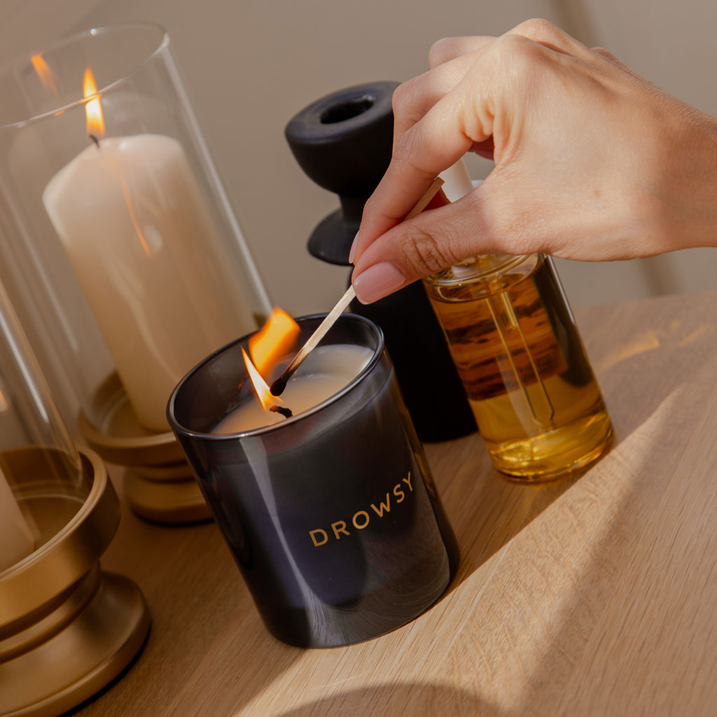 A hand lights the black Sleep Ritual Candle 220g with a match. Surrounding it are other candles and a bottle of oil on a wooden surface, all bathed in warm, natural light—ideal for a soothing bedtime ritual.