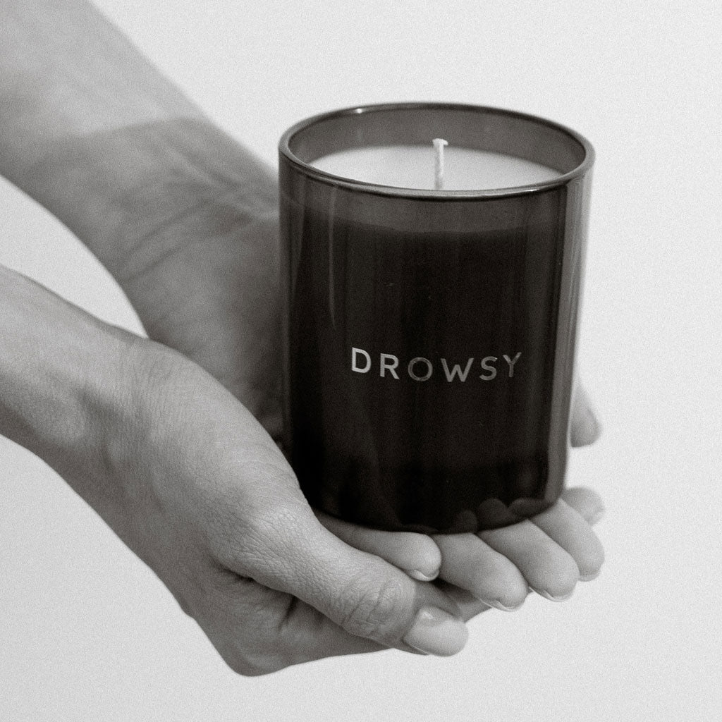 A person gently holds the Sleep Ritual Candle 220g in a dark glass jar with DROWSY on the label. The black and white image highlights hands cupping the candle—ideal for a soothing bedtime routine.