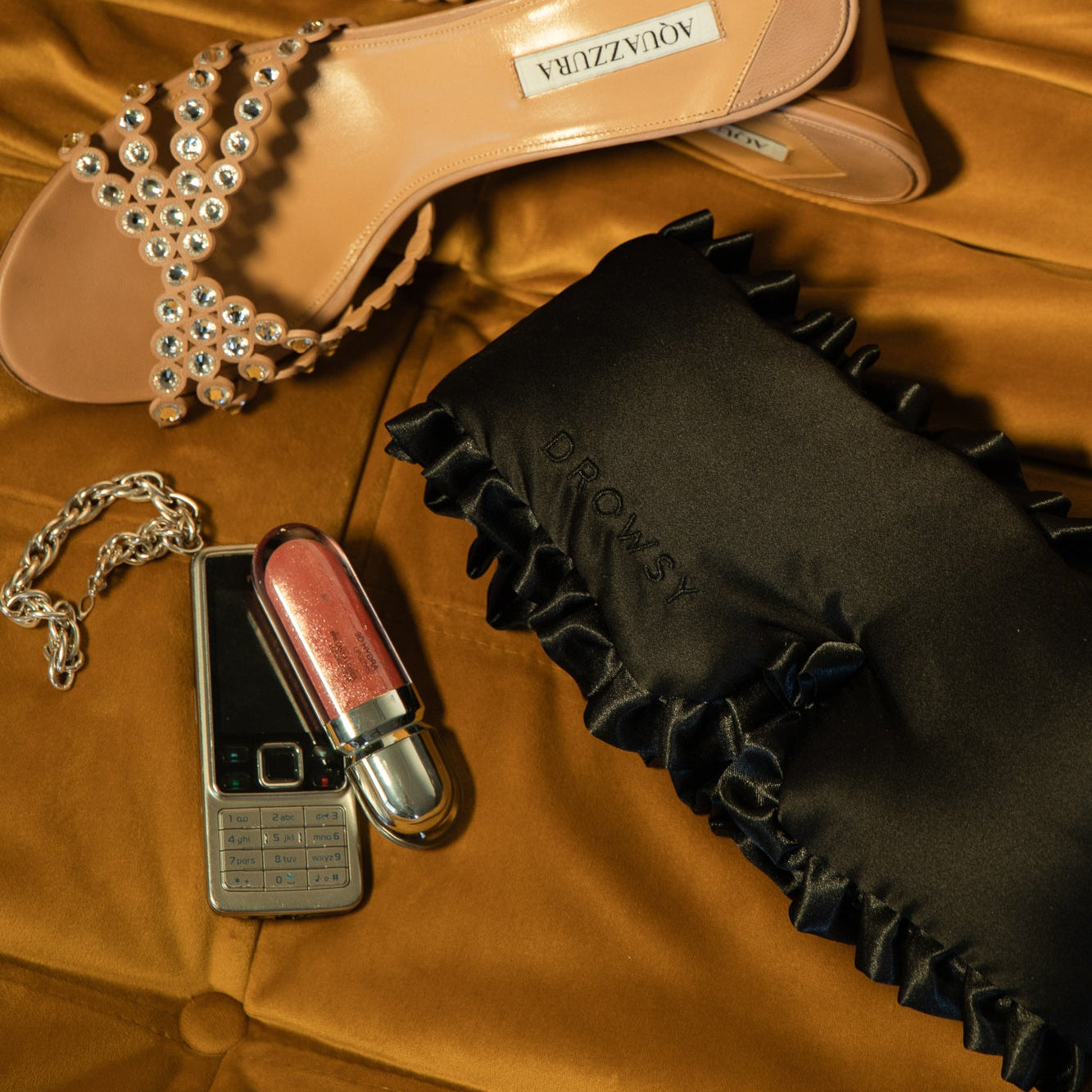 Sleep and the City is artfully arranged on a gold velvet surface alongside jeweled sandals, a mulberry silk sleep mask with DROWSY embroidery, a flip phone, silver chain, and pink lipstick for the ultimate beauty sleep vibe.