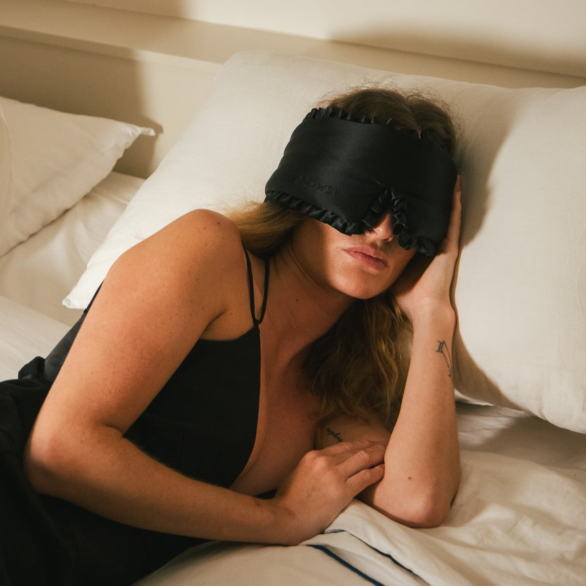 A woman with long hair lies on a bed, wearing a black sleep mask and black sleepwear. She is resting on white pillows, with one hand touching the side of her head.