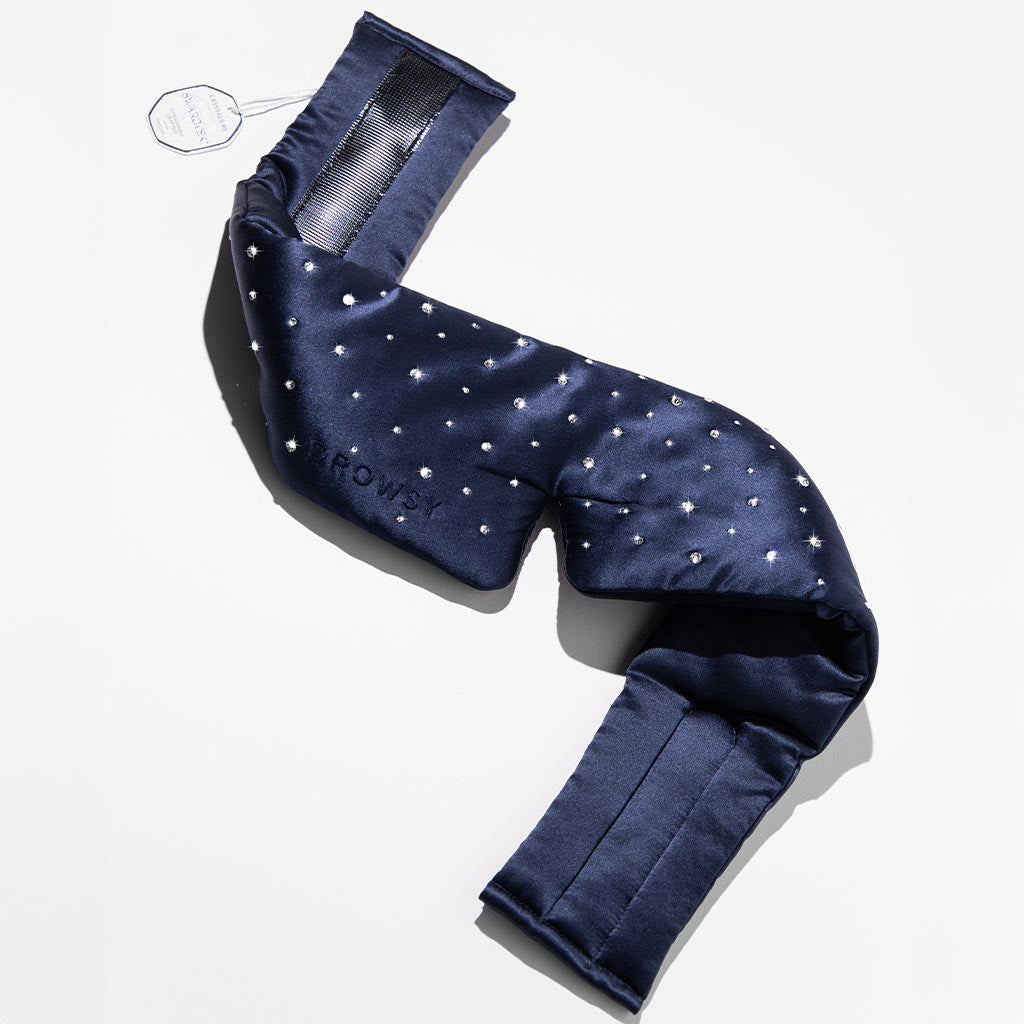 The Drowsy Silk Eye Mask Crystals by Swarovski® is shown on a white surface, featuring dark blue silk with sparkling crystal details, a satin finish, wide padded sides, and a tag for luxurious comfort and style.