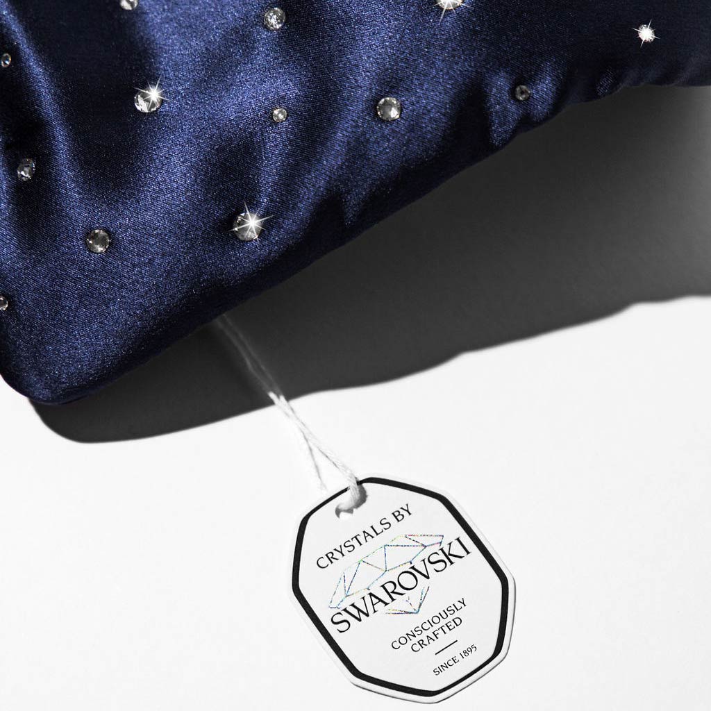 The Drowsy Silk Eye Mask Crystals by Swarovski® in navy blue silk features sparkling Swarovski crystals and a white tag reading Crystals by Swarovski, consciously crafted since 1895, attached with a white string on a white background.