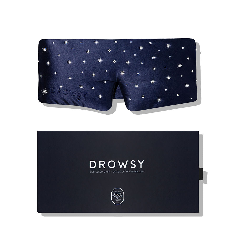 A dark blue silk eye mask embellished with Swarovski® crystals is displayed above a navy box labeled Drowsy Silk Eye Mask Crystals by Swarovski®, both set against a white background.