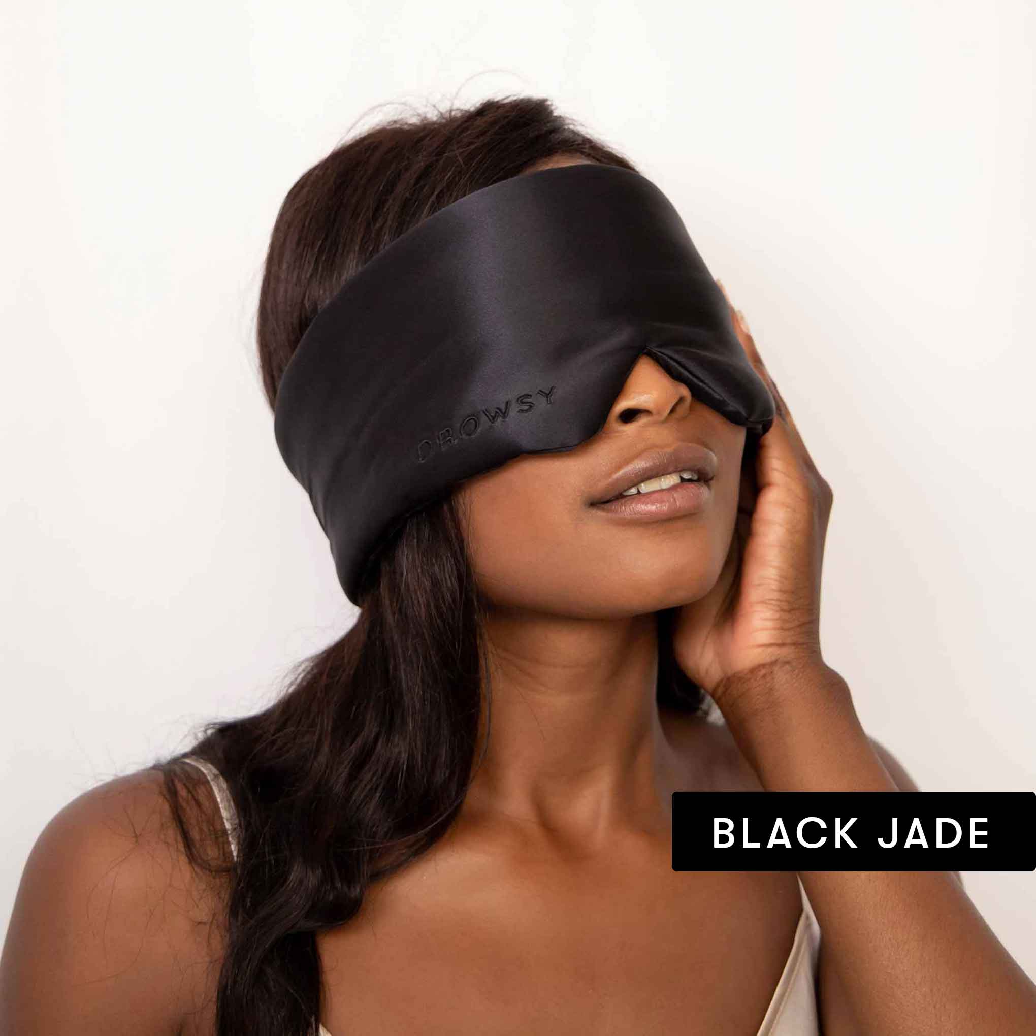 A woman with long dark hair wears a large black silky sleep mask labeled DROWSY from the Lovers Pack collection. A BLACK JADE tag appears at the bottom right as she lightly touches her face.