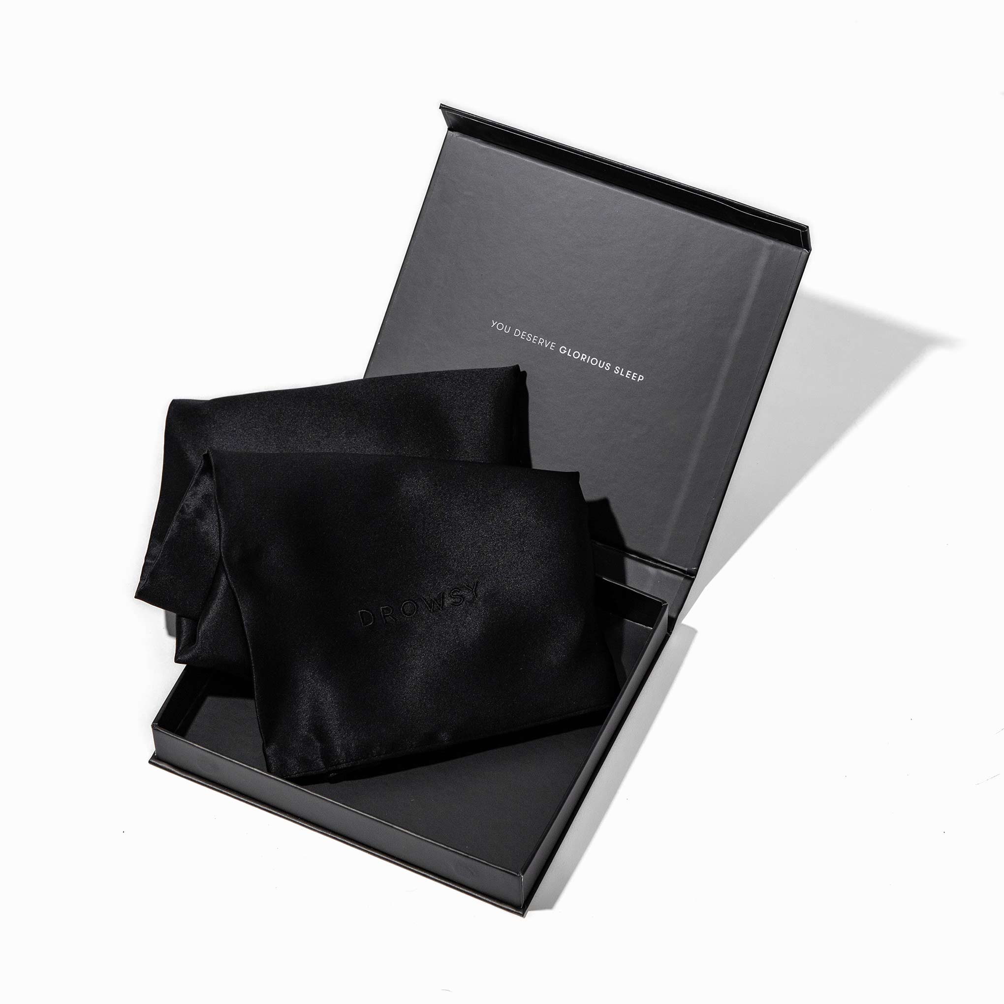 A black gift box with an open lid displays two folded Black Jade Silk Pillowcases—skincare and beauty essentials. The lid reads YOU DESERVE, GLORIOUS SLEEP, showcasing these hair-protecting silk pillowcases.