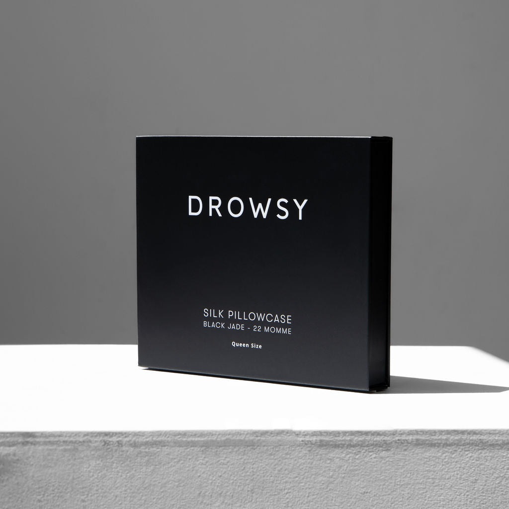 A black box labeled Black Jade Silk Pillowcase - 22 Momme, Queen Size stands upright on a white surface, the ideal beauty essential for protecting hair and skin, set against a neutral gray background.