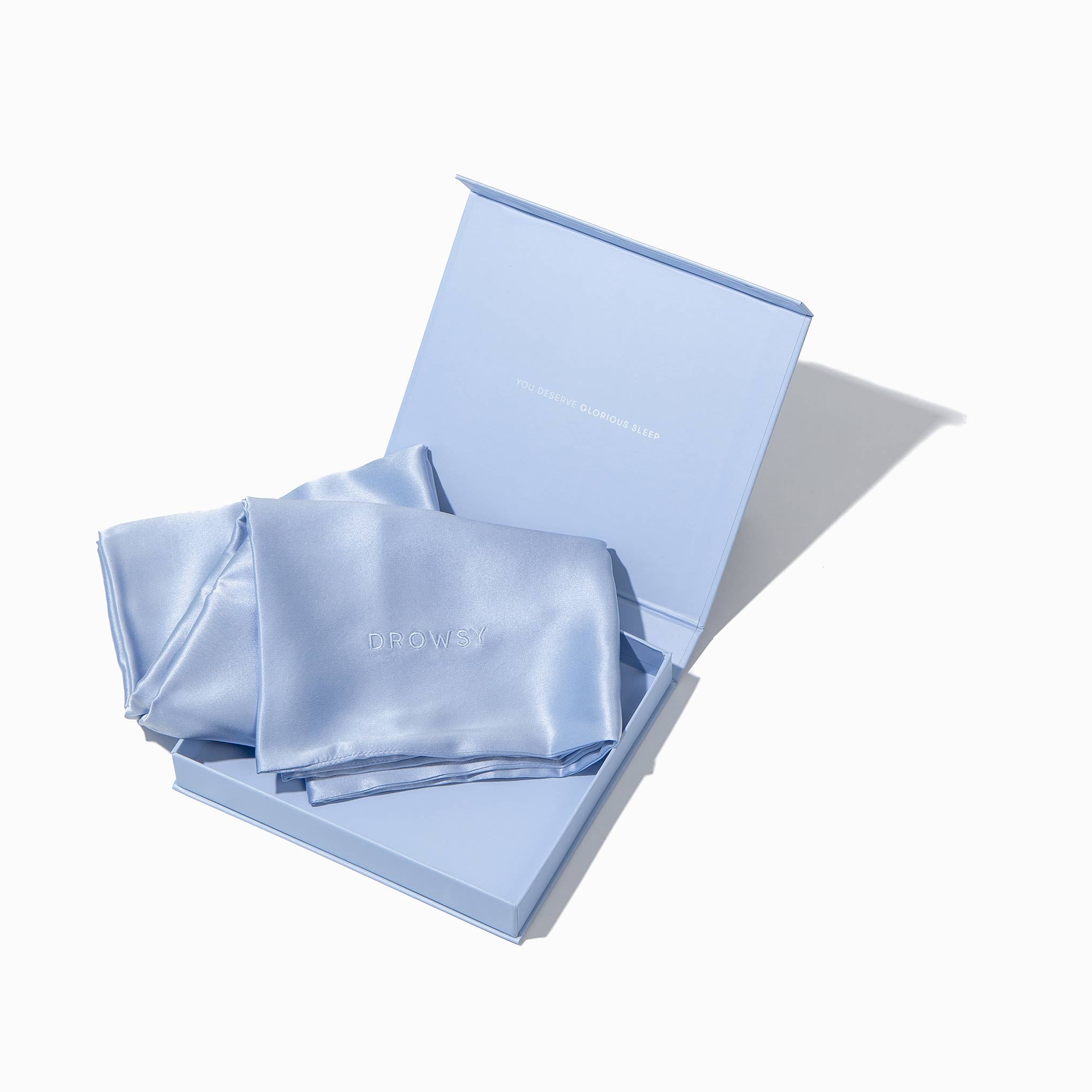 A light blue Blue Belle Silk Pillowcase, draped over the edge of an open matching blue gift box on a white background—a hair protecting, skincare and beauty essential.