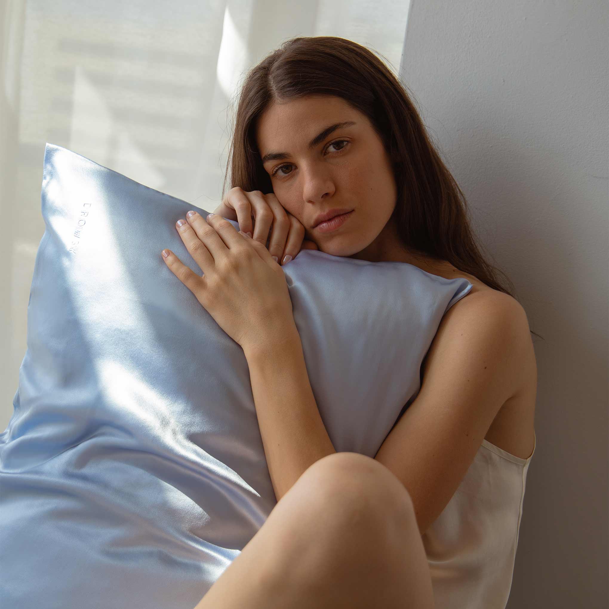 A woman with long brown hair leans against a white wall, thoughtfully holding the Blue Belle Silk Pillowcase—ideal for hair protection and beauty benefits—as soft natural light streams in through a nearby window.