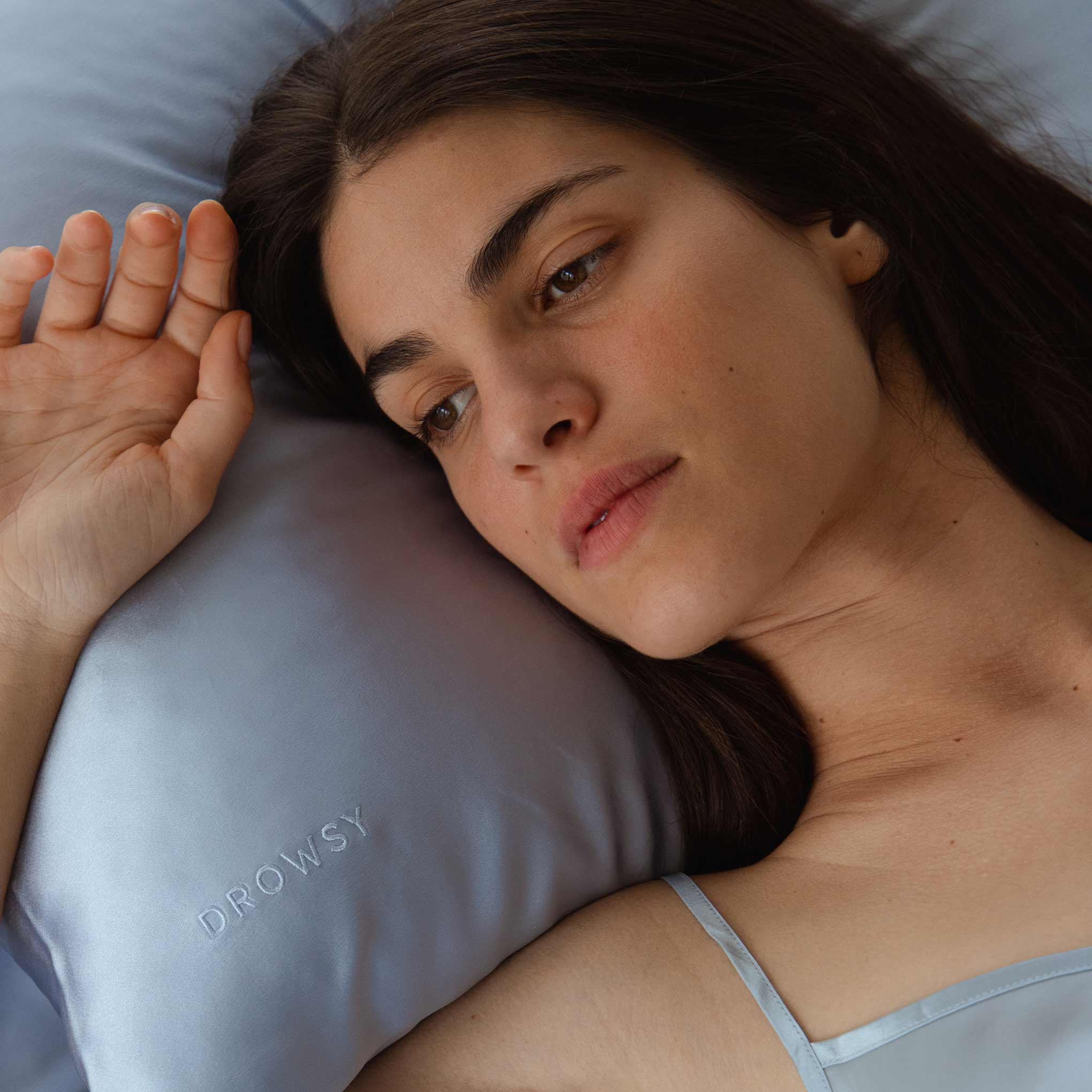 A woman with long brown hair lies peacefully on the Blue Belle Silk Pillowcase, embroidered with DROWSY. She looks relaxed in a light blue camisole—an ideal hair-protecting, skincare, and beauty essential.