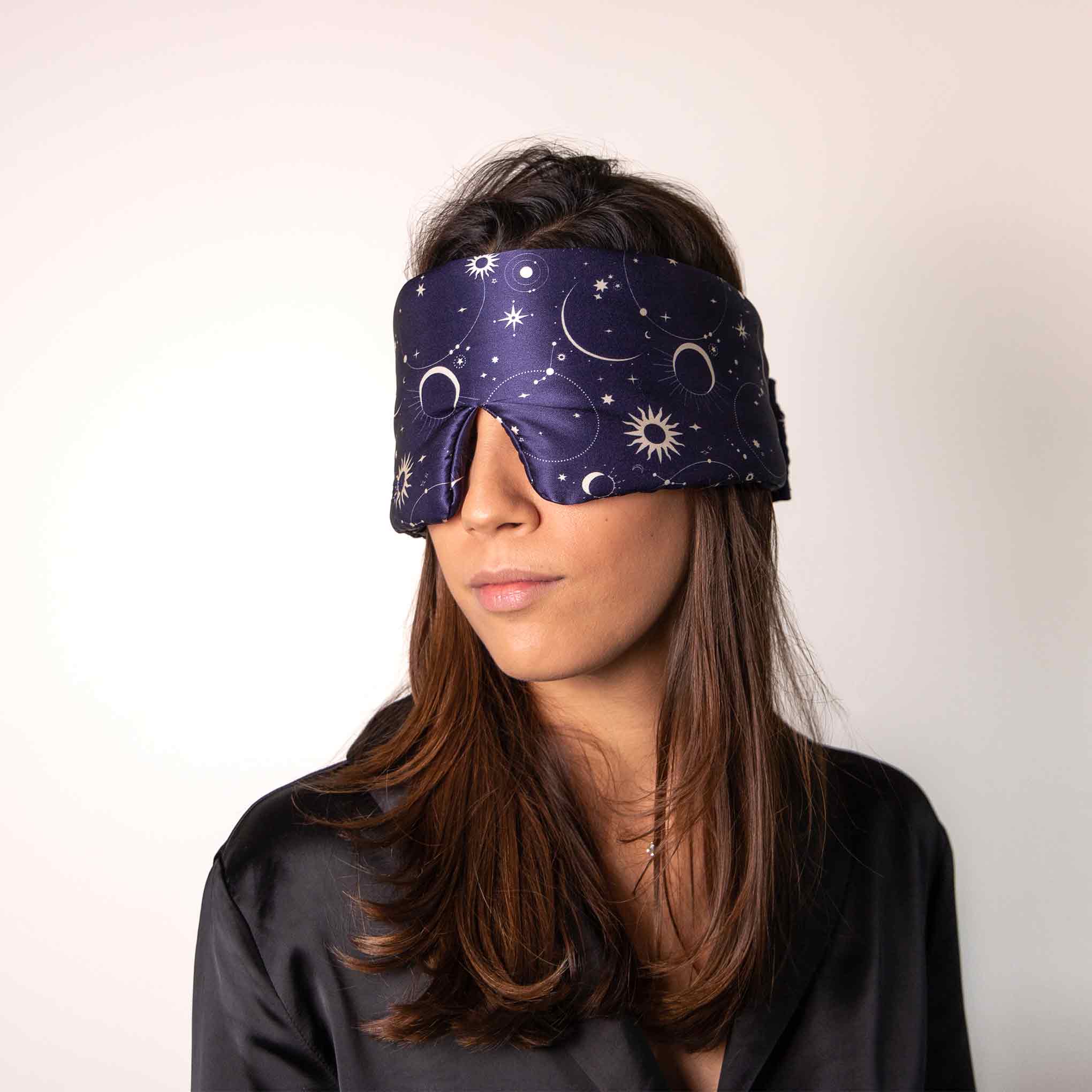 A woman with long brown hair wears a navy blue sleep mask decorated with stars, moons, and suns. She is dressed in a black satin top and stands against a plain, light background.