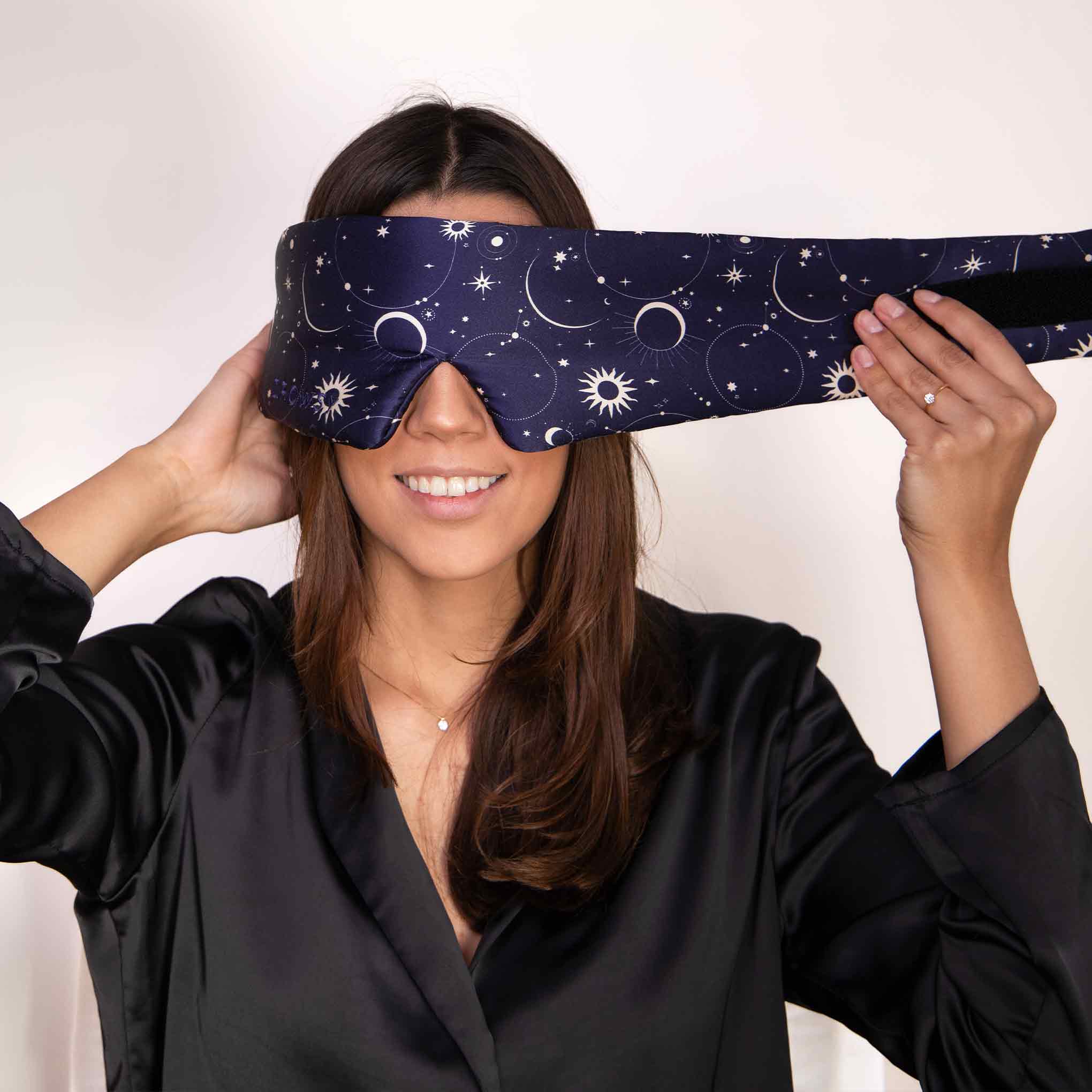 Smiling woman wearing a dark sleep mask with moon and star patterns, holding the sides as she puts it on, dressed in a black robe against a plain background.