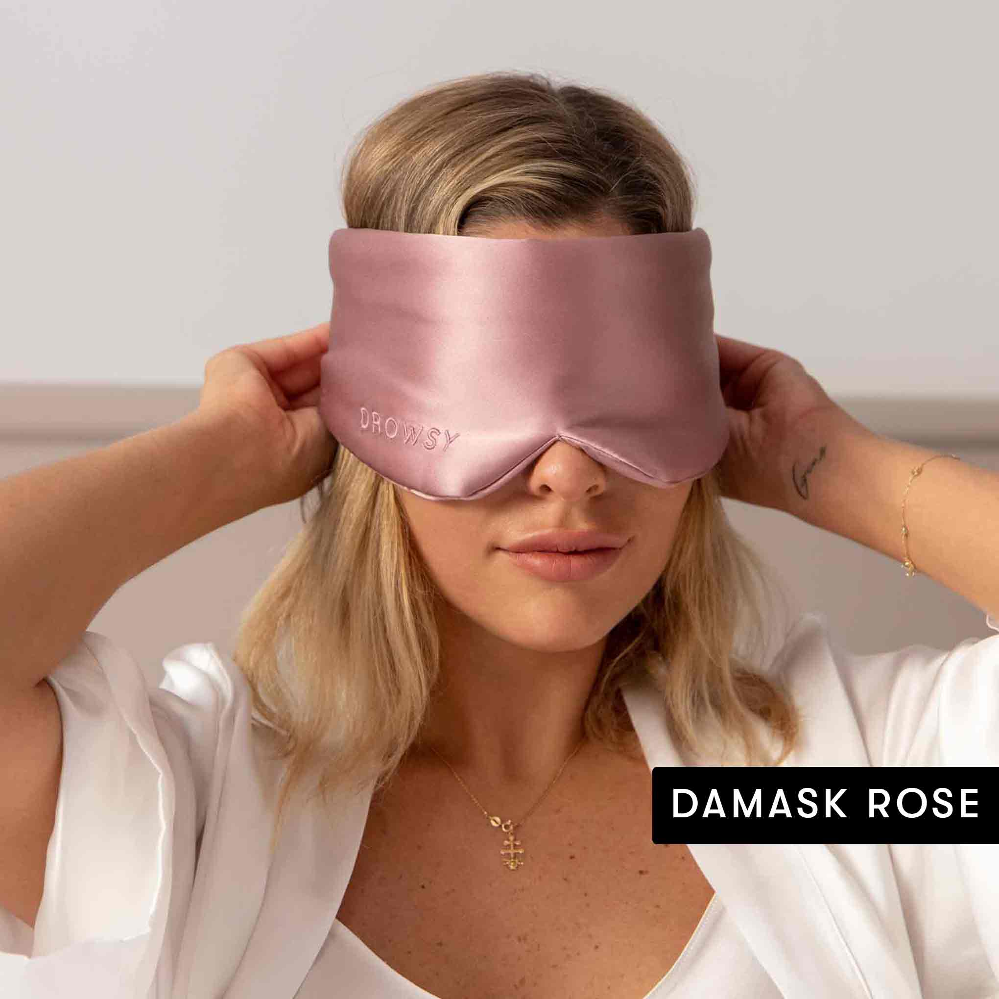 A woman in a white robe with loose blonde hair adjusts a silky pink sleep mask labeled Family Pack over her eyes. The text DAMASK ROSE appears in the lower right corner.