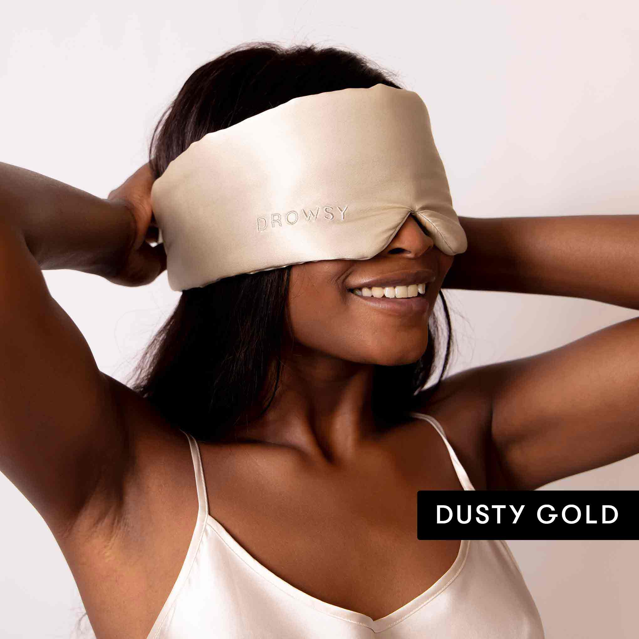 A woman smiles as she adjusts a light gold satin sleep mask embroidered with DROWSY from the Besties Pack. The text DUSTY GOLD appears in the bottom right corner.