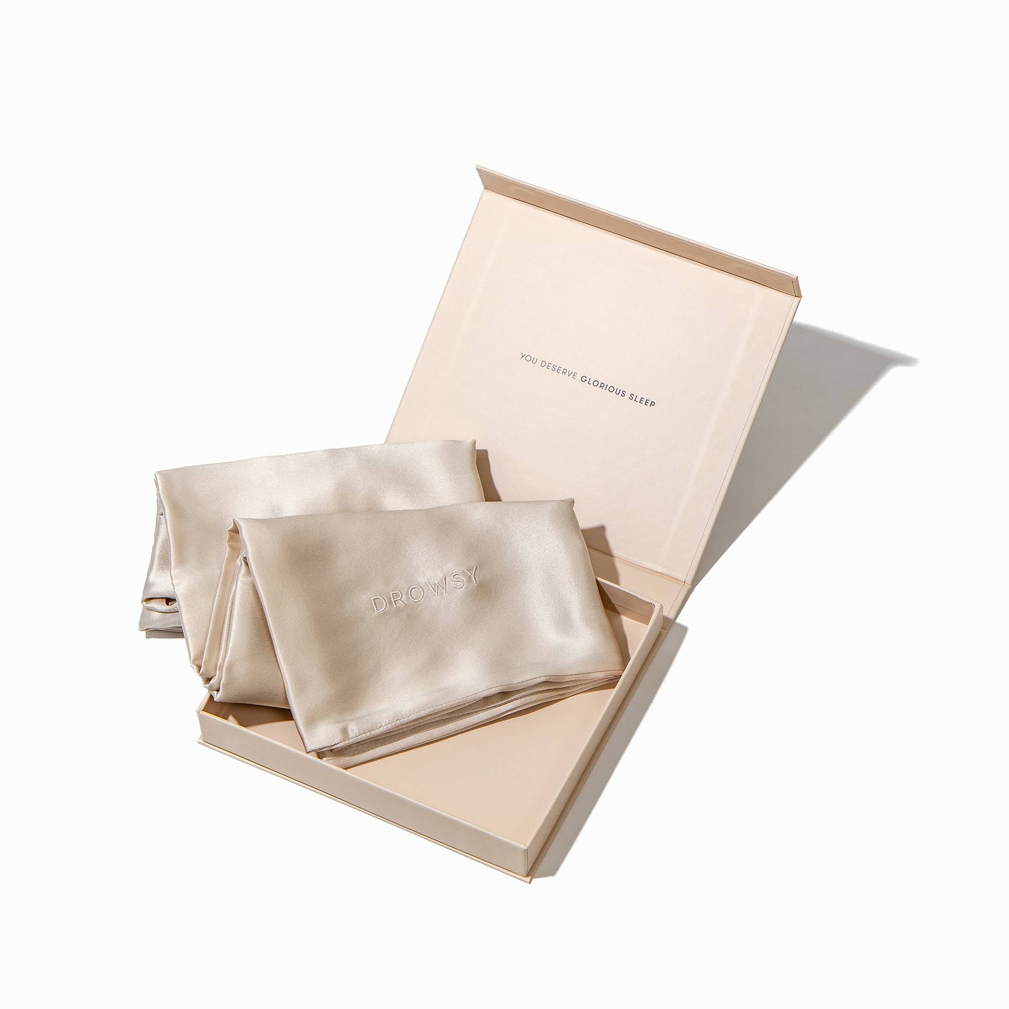 The Dusty Gold Silk Pillowcase set, partly folded beside an open box reading YOU DESERVE GLORIOUS SLEEP inside the lid, features a subtle embroidered logo and is a true skincare essential.