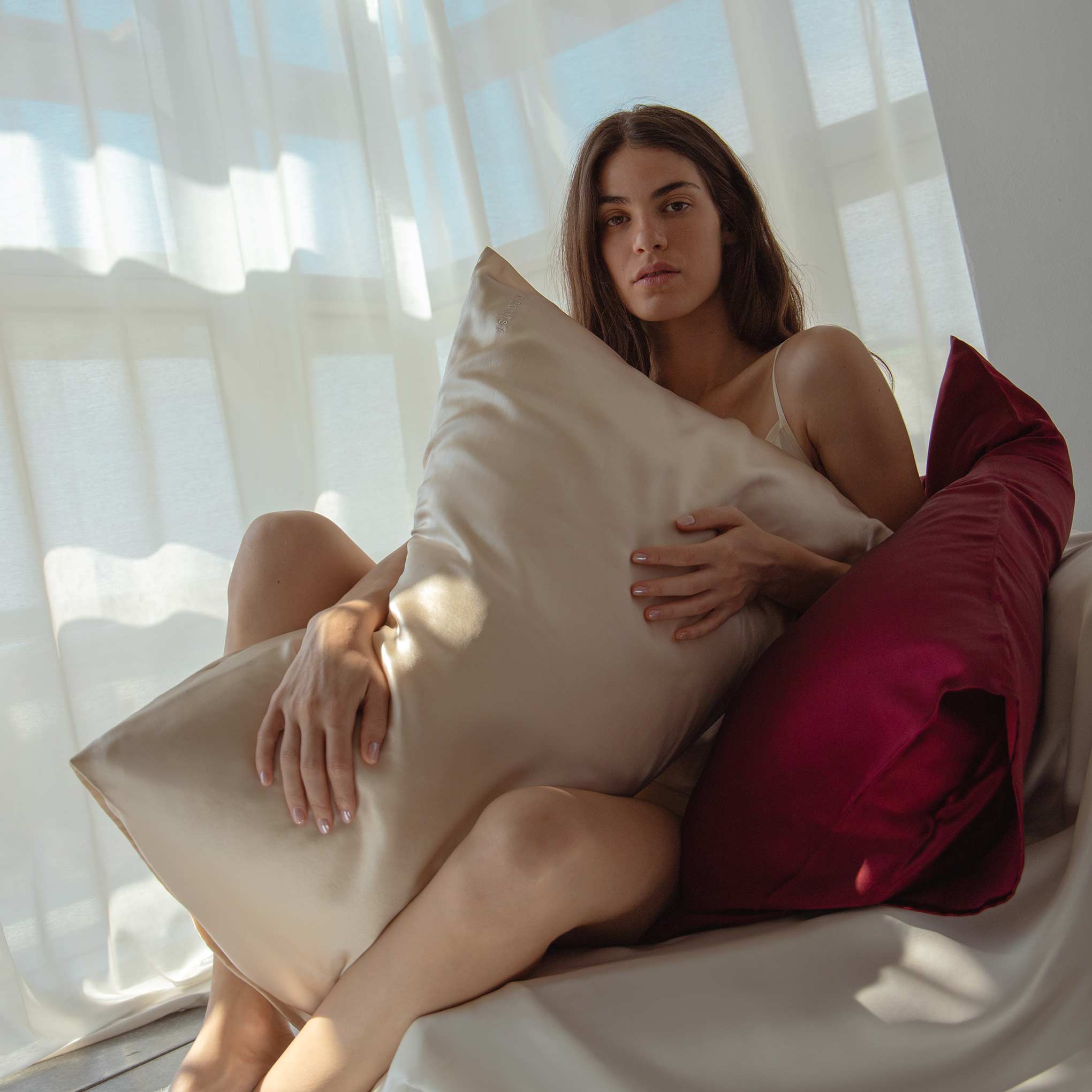 A woman with long hair sits on a couch by a window, holding a Dusty Gold Silk Pillowcase—an essential for hair protection and skincare. Soft sunlight filters through sheer curtains in the background.
