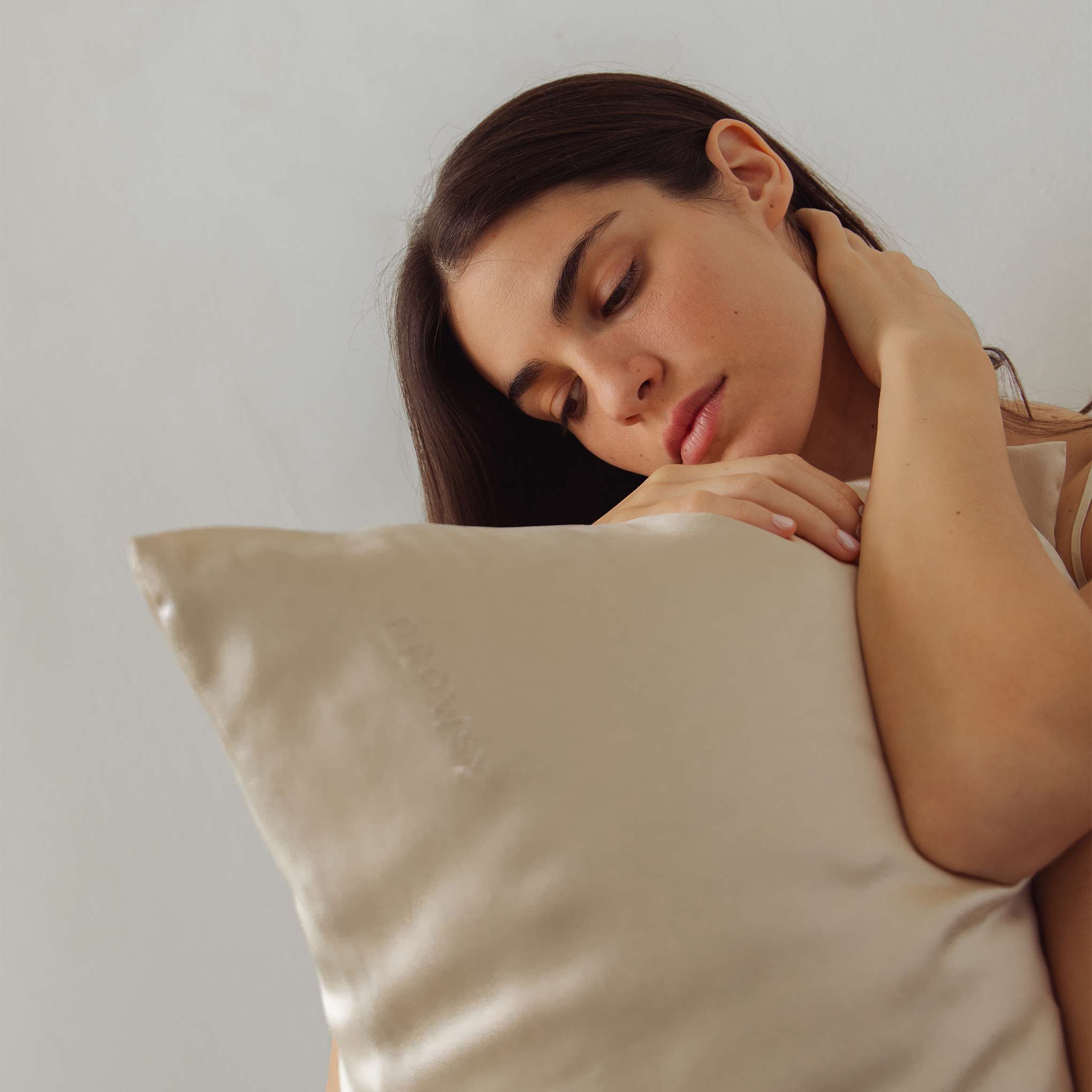 A woman with long dark hair hugs the Dusty Gold Silk Pillowcase, resting her head on it and looking down serenely—highlighting this hair-protecting essential against a plain background.