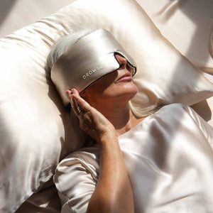 A woman with gray hair lies on a Dusty Gold Silk Pillowcase, wearing a large, light-colored DROWSY sleep mask. Dressed in shiny satin, she gently touches the mask—showcasing her favorite skincare essential.