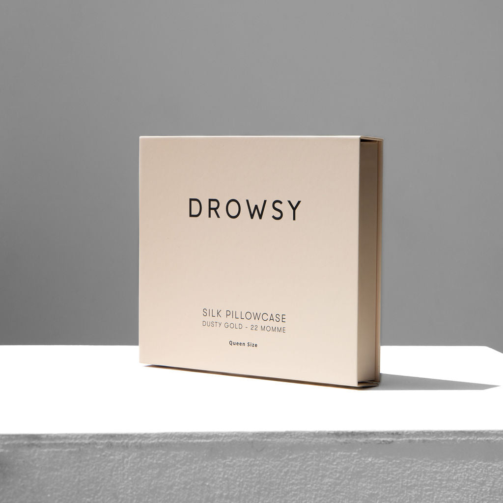 A beige box labeled Dusty Gold Silk Pillowcase sits on a white surface against a gray background, showcasing this silk pillowcase as a skincare essential and hair protector.