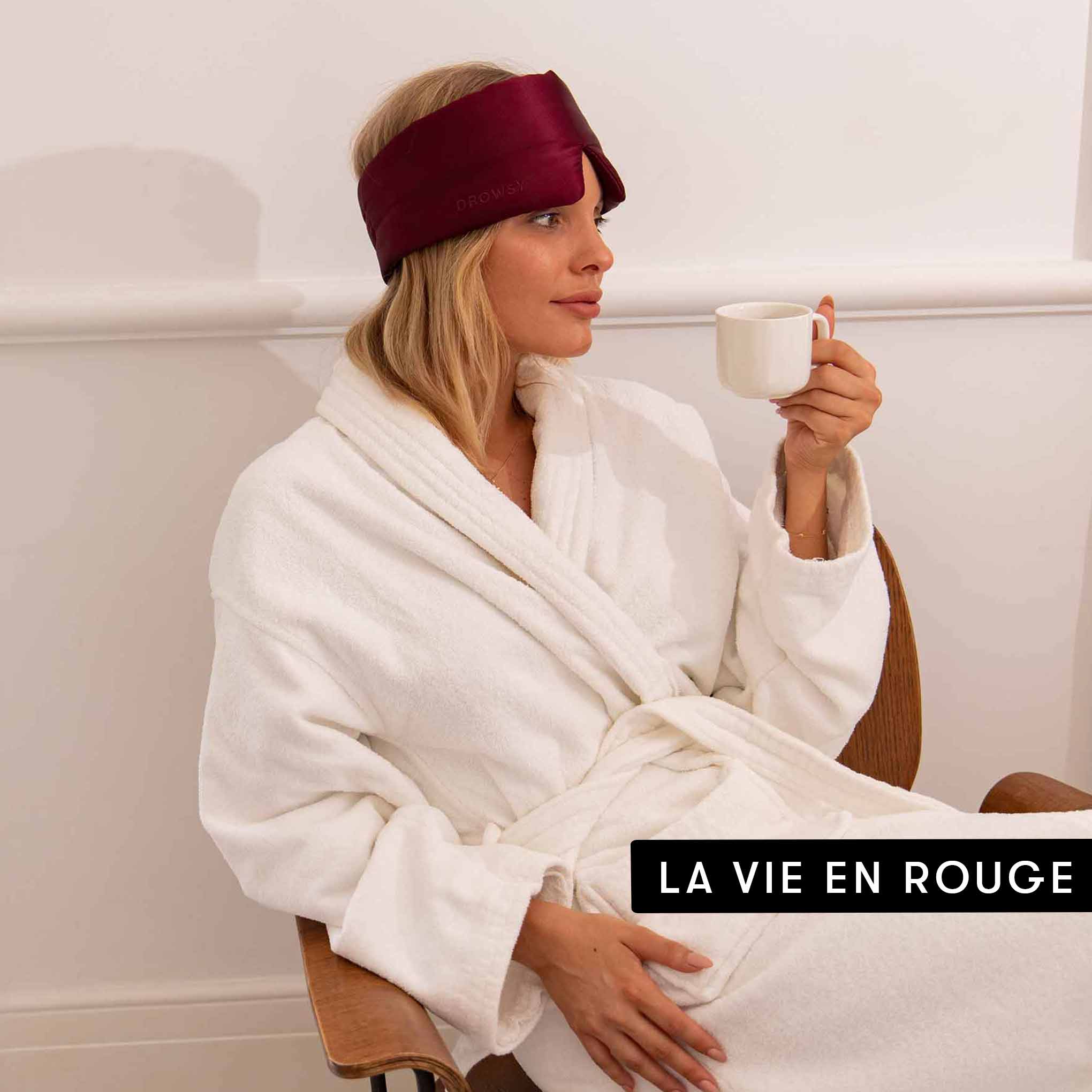A woman in a white bathrobe sits in a chair, wearing a burgundy Lovers Pack sleep mask pushed up on her head and holding a white mug. The text LA VIE EN ROUGE is at the bottom right.