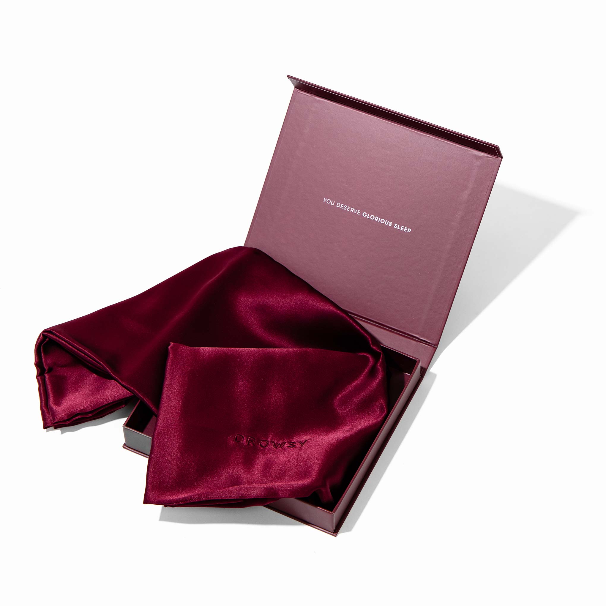 A luxurious burgundy silk sleep mask is shown in an open box beside the La Vie En Rouge Silk Pillowcase. This beauty essential has a subtle embroidered logo and a lid reading YOU DESERVE GLORIOUS SLEEP for hair-protecting rest.