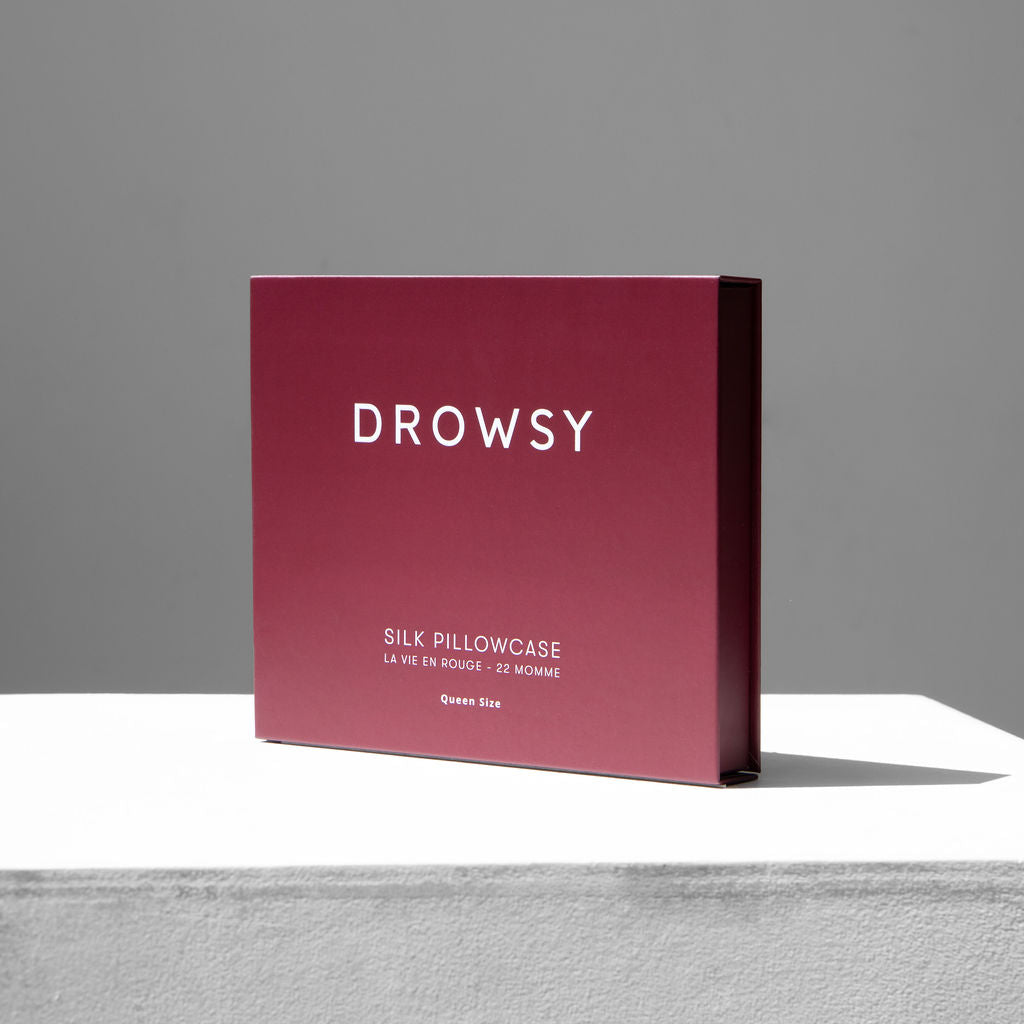 A maroon La Vie En Rouge Silk Pillowcase box labeled "Drowsy" with "22 Momme, Queen Size" text is displayed on a white surface against a gray background, highlighting this silk pillowcase as a skincare and beauty essential.