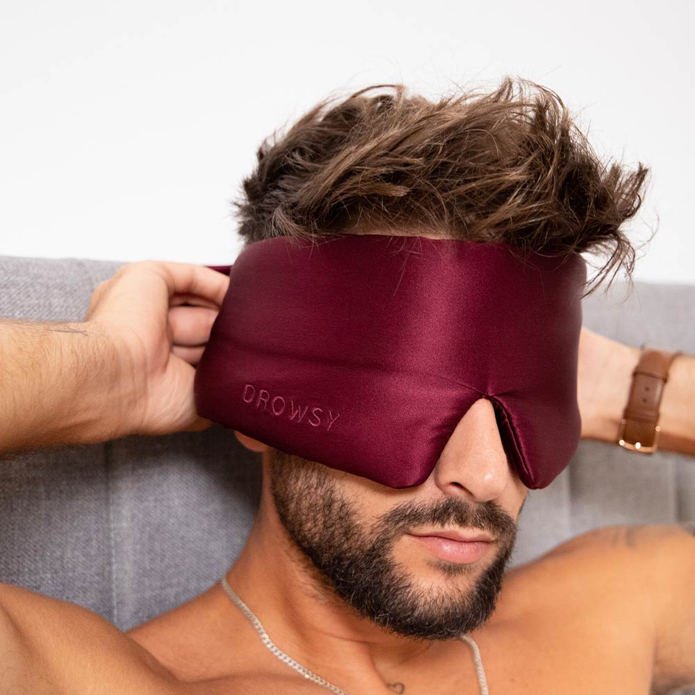 A man with tousled hair adjusts a large, burgundy silk La Vie En Rouge eye mask while sitting up in bed against a gray headboard, ready for the ultimate beauty sleep.
