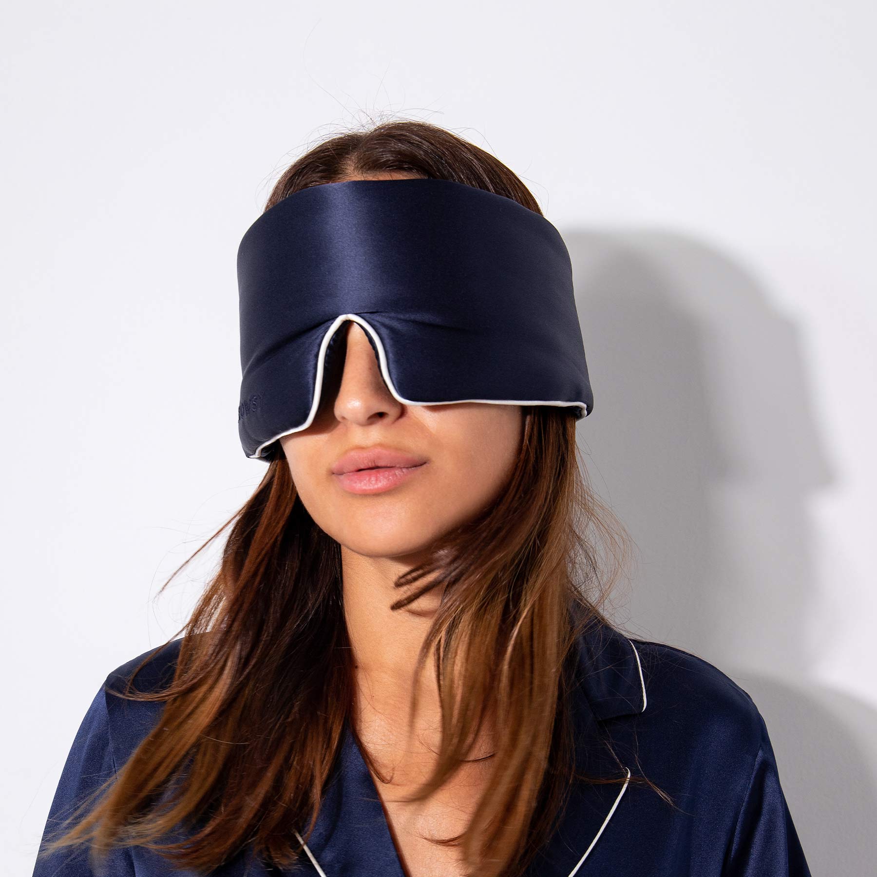 A woman wears the Midnight Blue - Pipe Dreams Edition silk sleep mask and matching pajamas, standing against a white background with her hair partly visible under the mask.