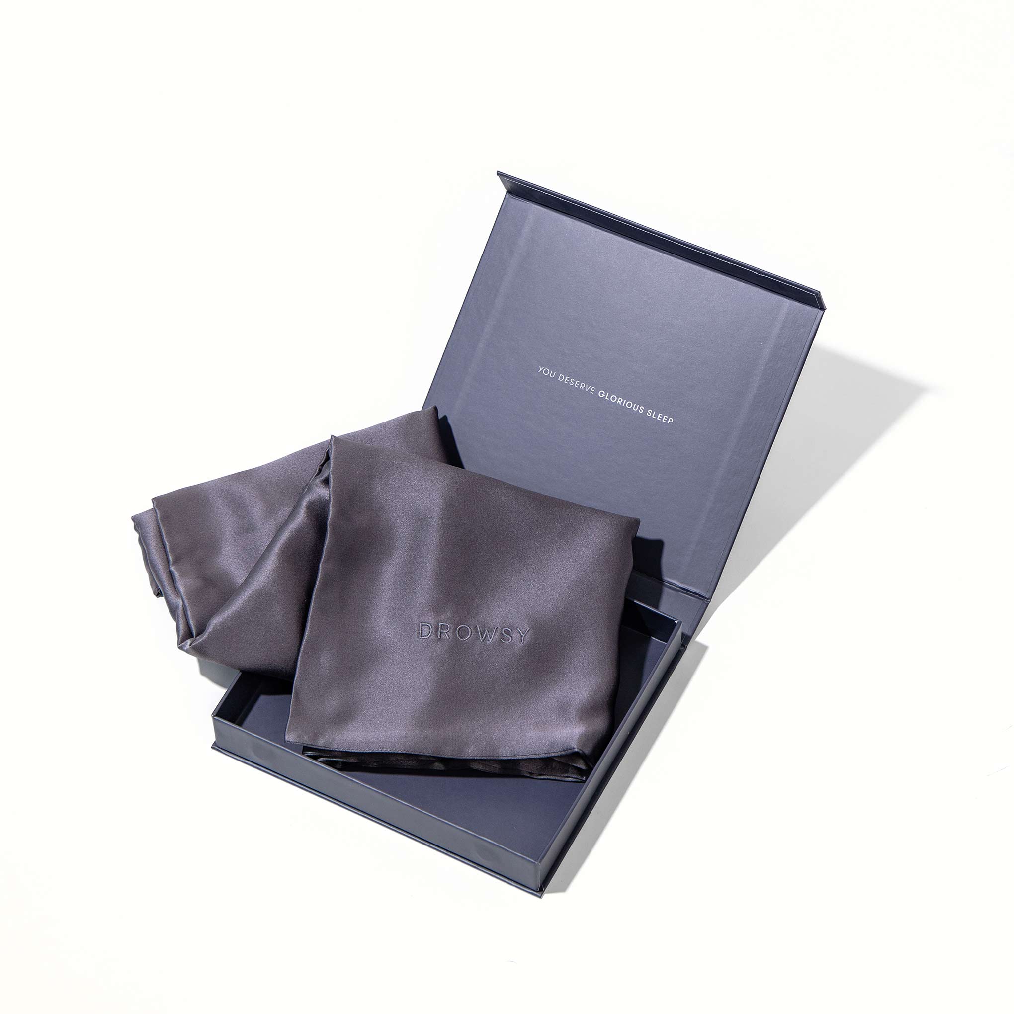 The Moonlight Shadow Silk Pillowcase in dark grey, embossed with PROWS, is partially folded in an open, matching dark blue gift box with text inside the lid, all set against a white background. Perfect for skincare and hair protection.