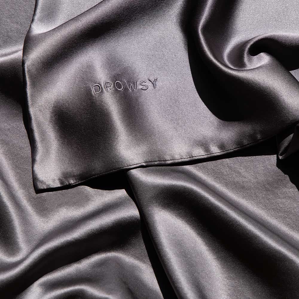 A close-up of smooth, shiny black silk fabric with the word DROWSY embroidered on one corner, showcasing the luxurious texture and sheen of the material.