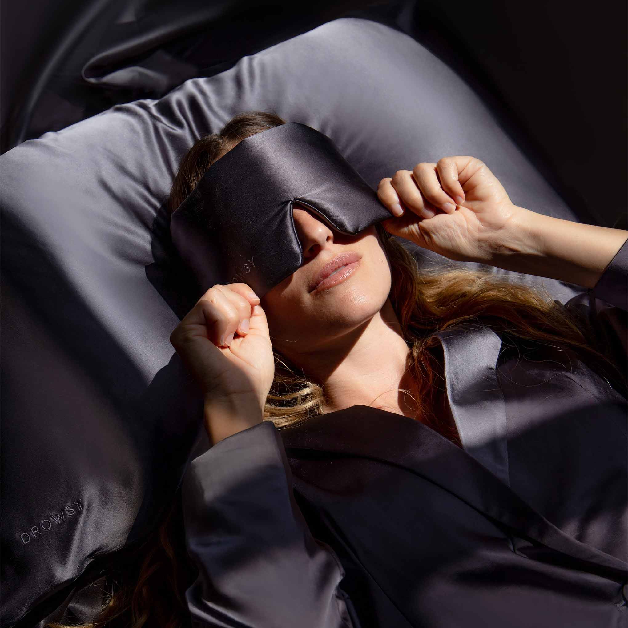 A woman relaxes on a bed with gray satin sheets, wearing an eye mask and pajamas, as sunlight highlights her face. She embraces her hair-care ritual with the Moonlight Shadow Silk Pillowcase.