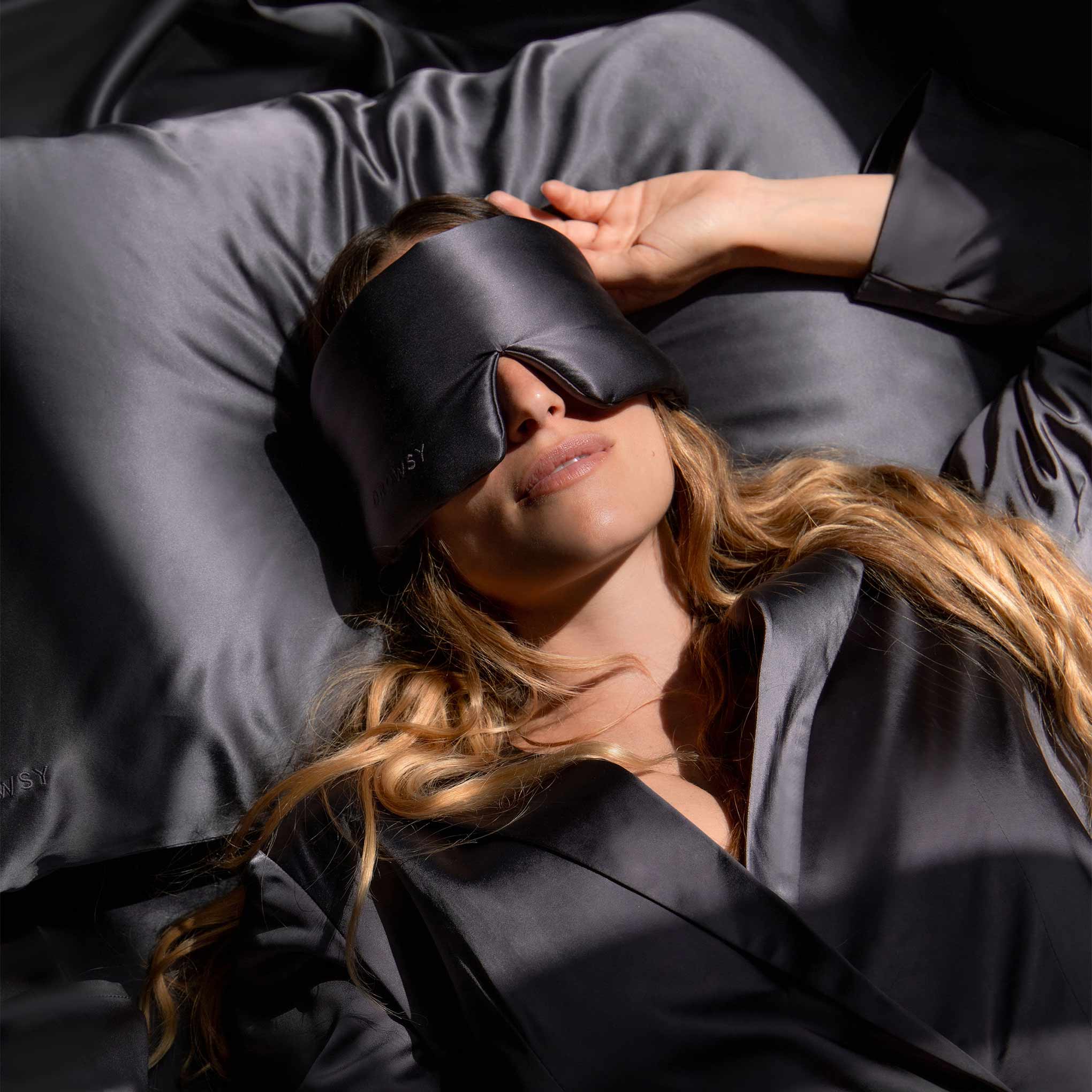 A woman with long, wavy hair rests peacefully on a dark bed, wearing matching satin pajamas and a sleep mask, her hand gently touching her forehead as she relaxes on the Moonlight Shadow Silk Pillowcase.